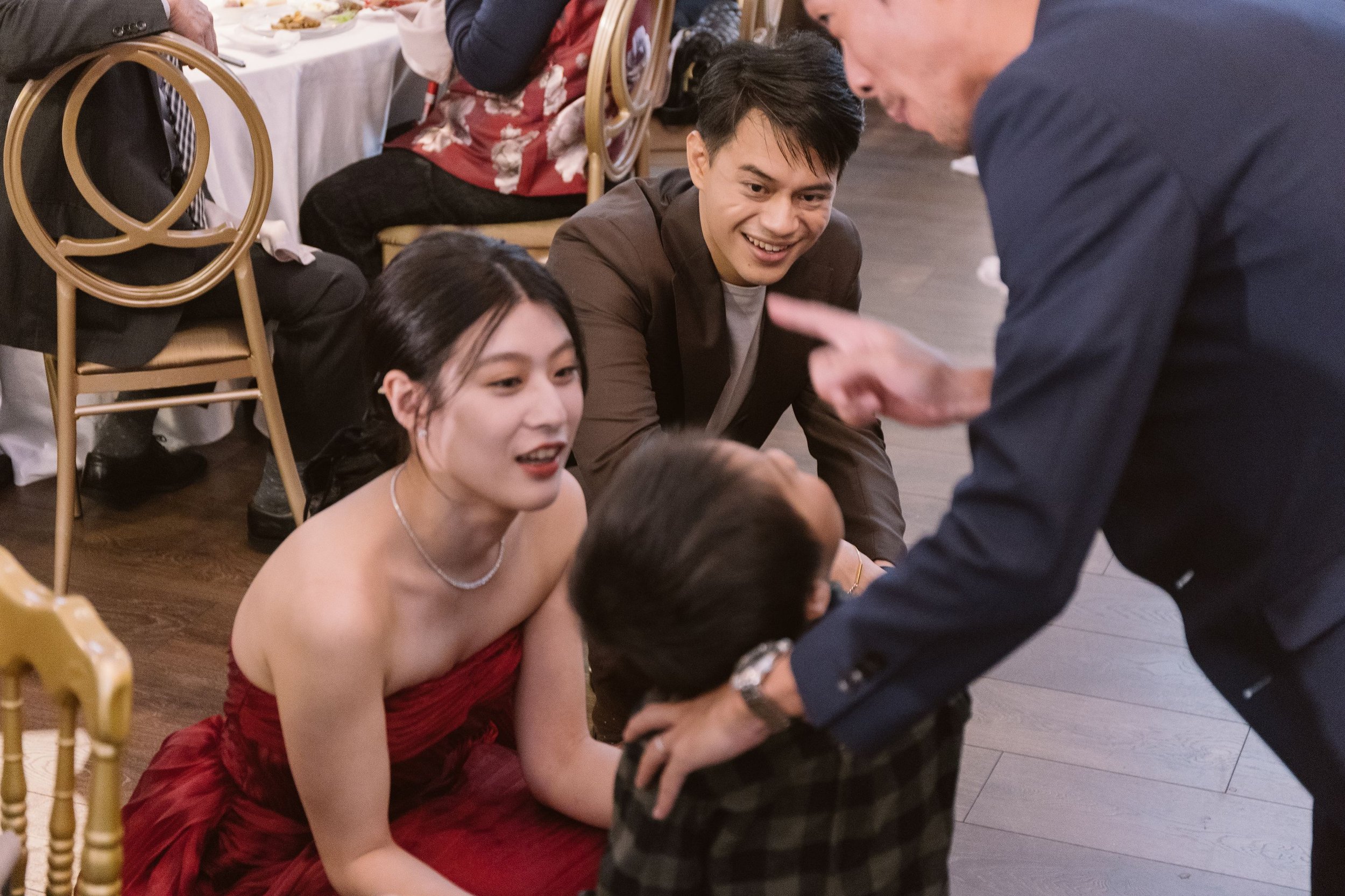 A young woman in a strapless red dress sitting on the floor, smiling as she interacts with a man in a suit. Another man with black hair and a brown blazer is also on the floor, smiling and looking engaged in conversation. The scene is inside a banque