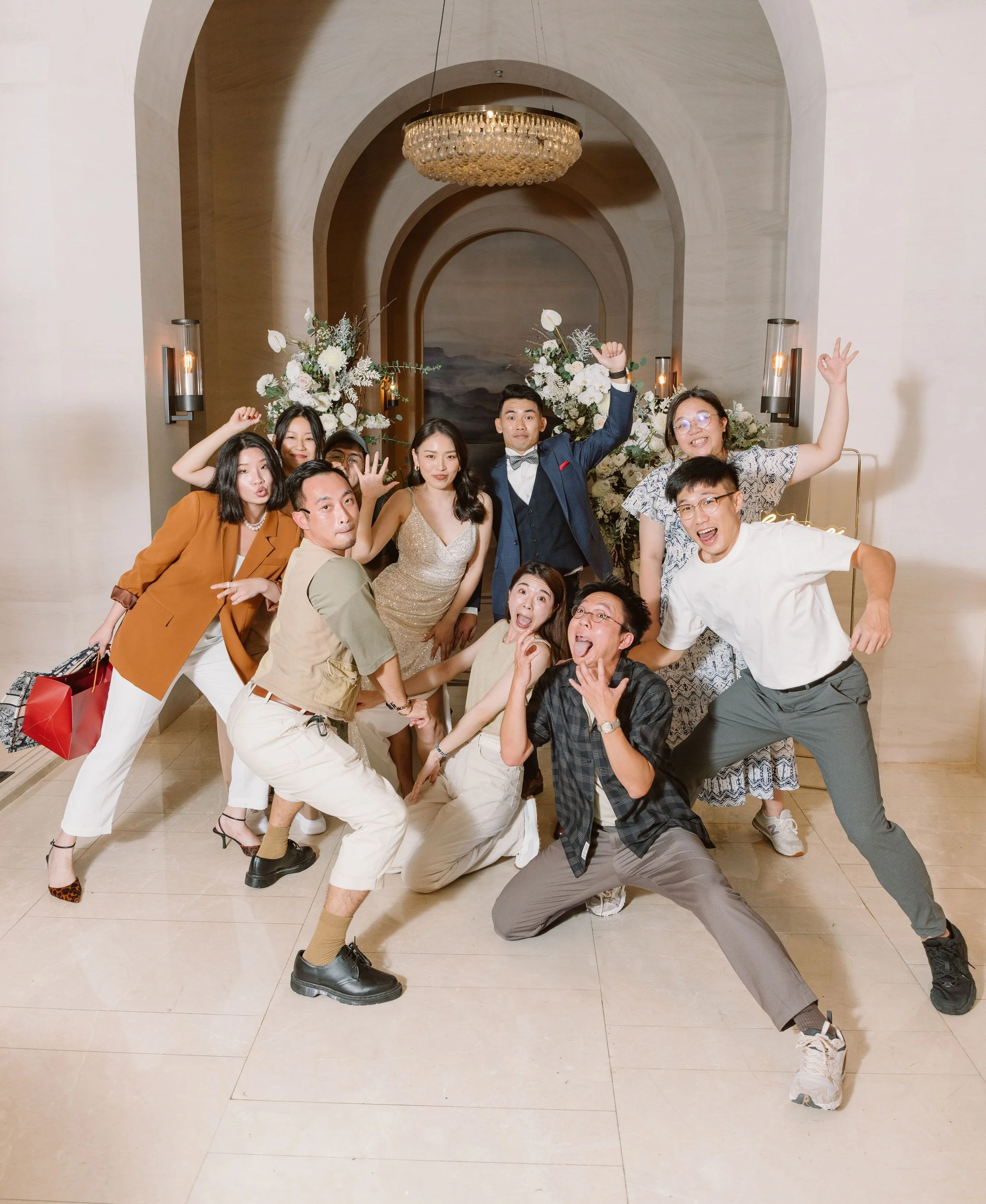 Group of people celebrating at a wedding or party, standing in front of floral arrangements and a chandelier in an elegant venue, with some making playful gestures and smiling.
