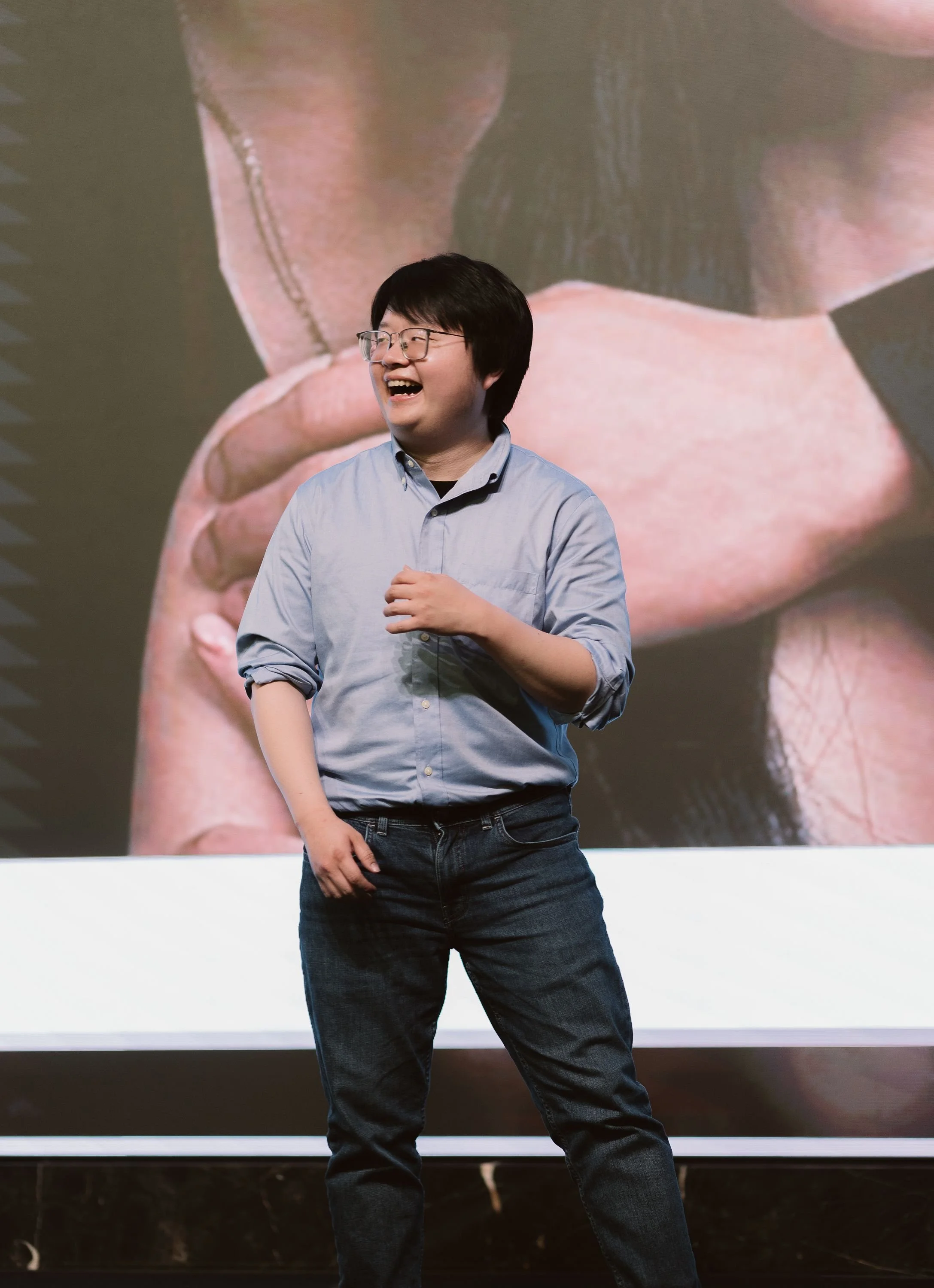 Man with black hair, glasses, in a light blue button-up shirt and dark jeans, smiling and standing in front of a large screen displaying a close-up image of a finger with a ring.