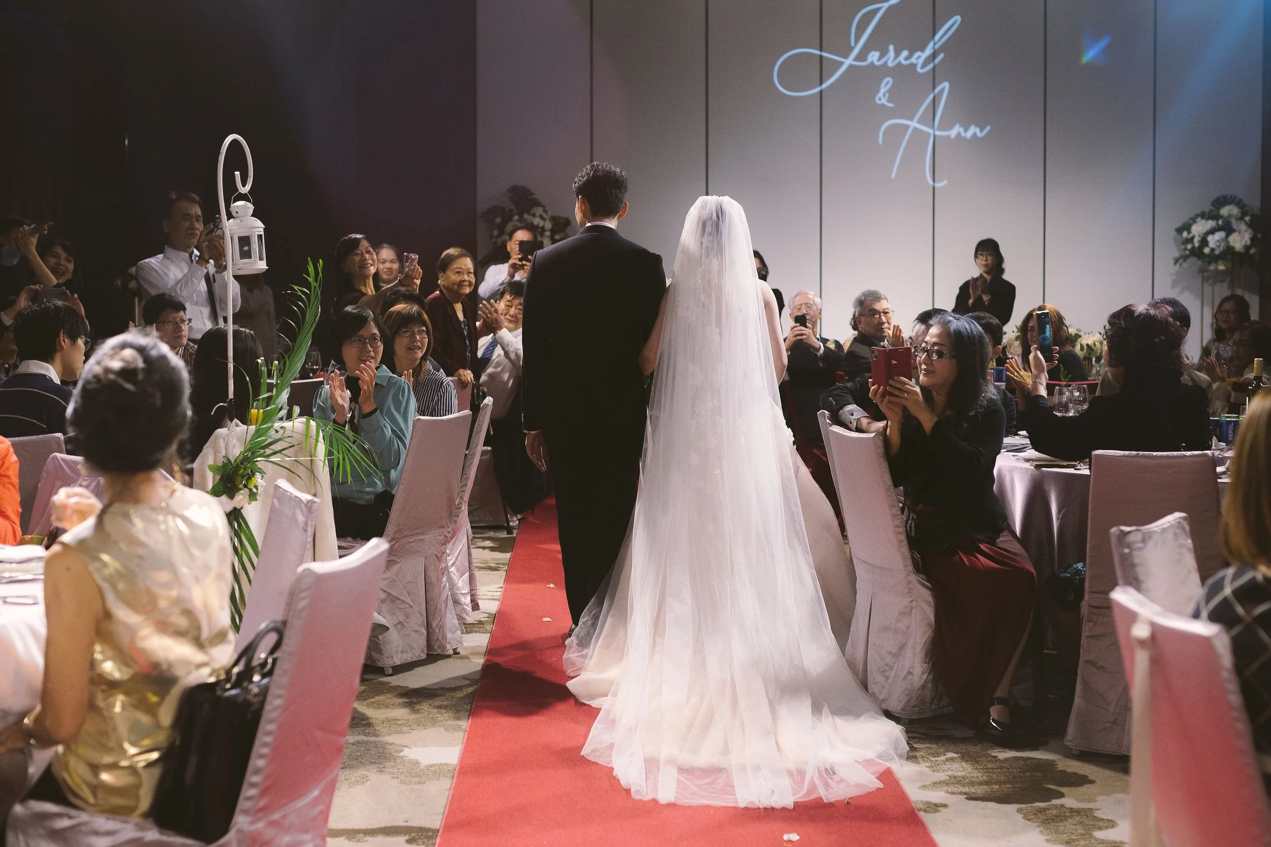 A wedding ceremony with a bride and groom standing on a red carpet, facing an audience in a banquet hall. The bride is wearing a white wedding gown with a veil, and the groom is in a black suit. Guests are seated at round tables, some taking photos a