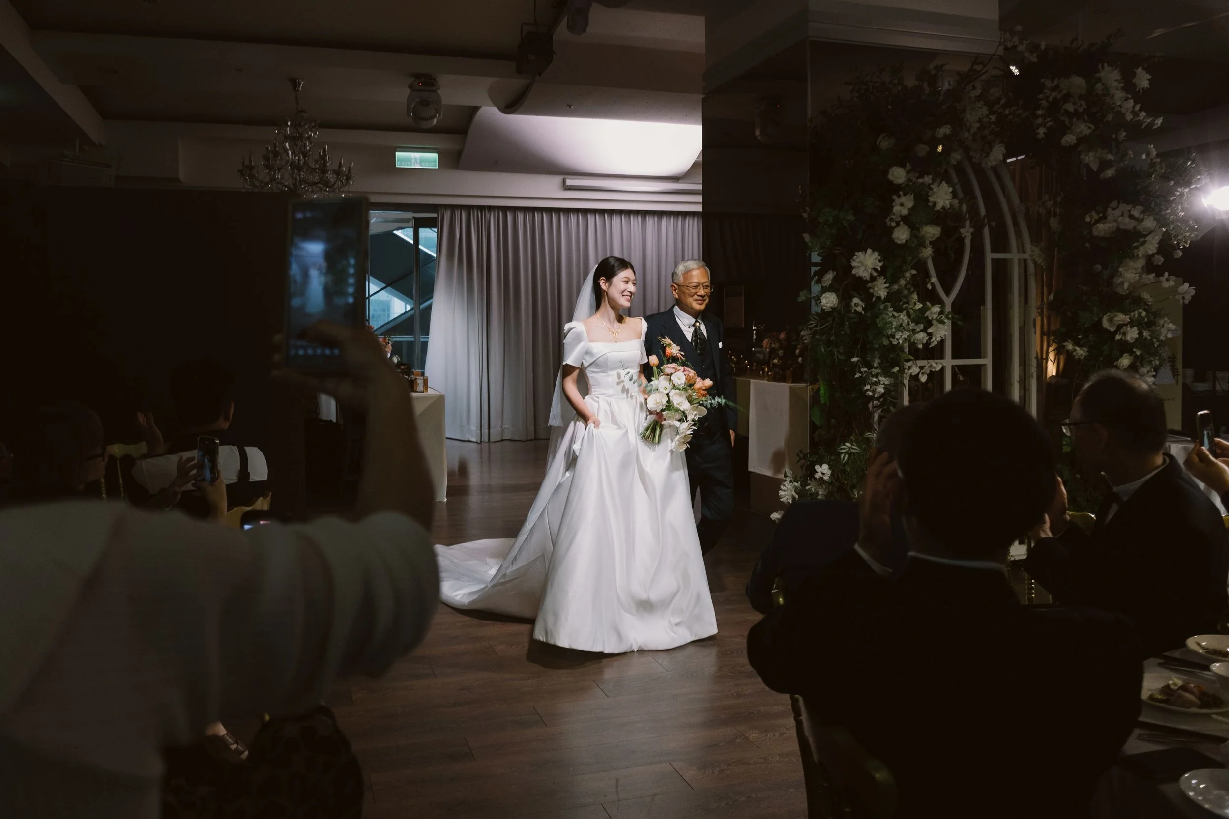A bride in a white wedding dress holding a bouquet of flowers walking down the aisle with an older man, likely her father, during a wedding ceremony. Guests are seated on either side taking photos.