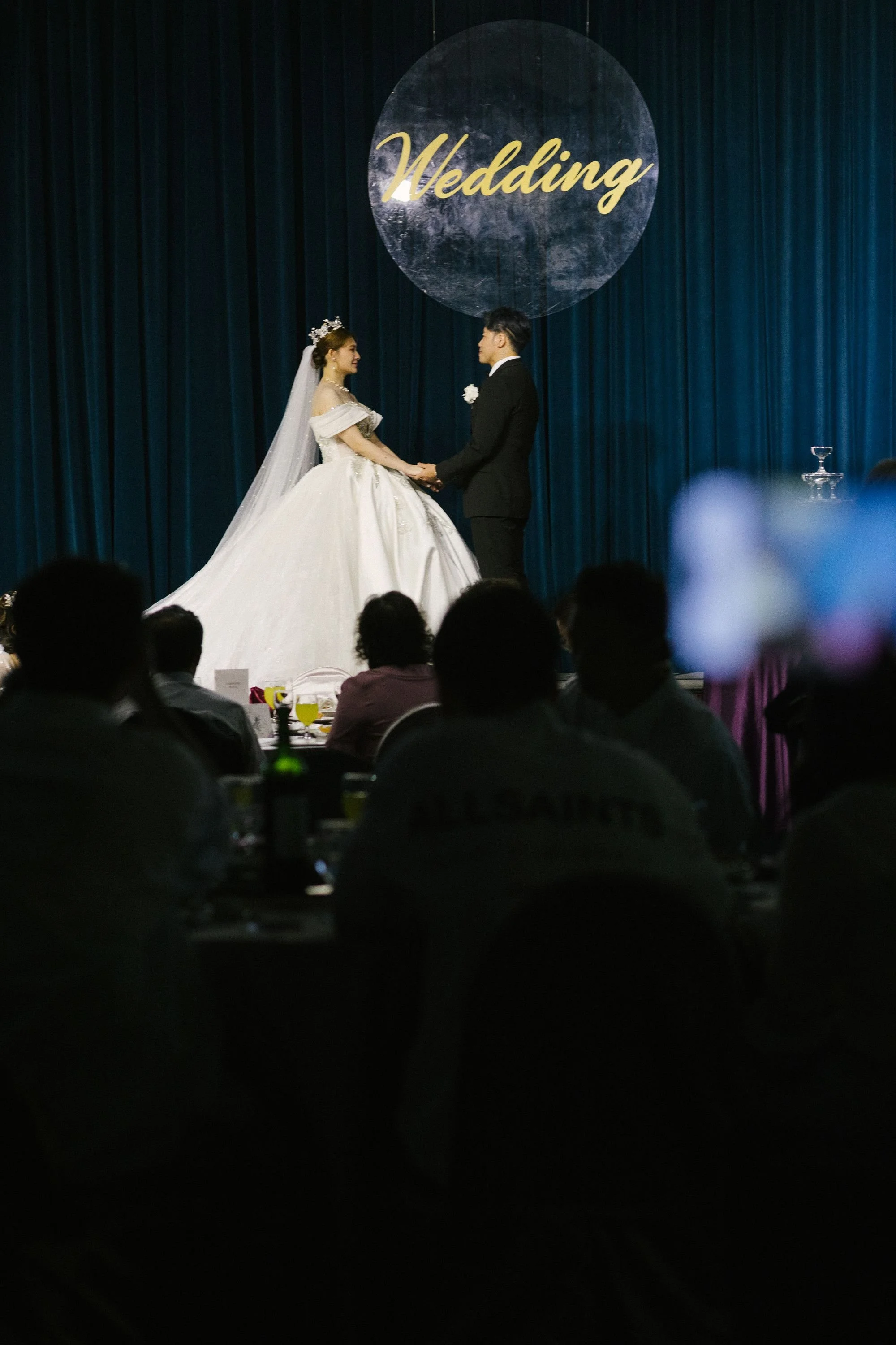 A bride and groom holding hands and looking at each other during a wedding ceremony on a stage with a dark blue curtain and a large circular sign reading 'Wedding' in the background. The bride is wearing a white gown and a tiara, and the groom is in 