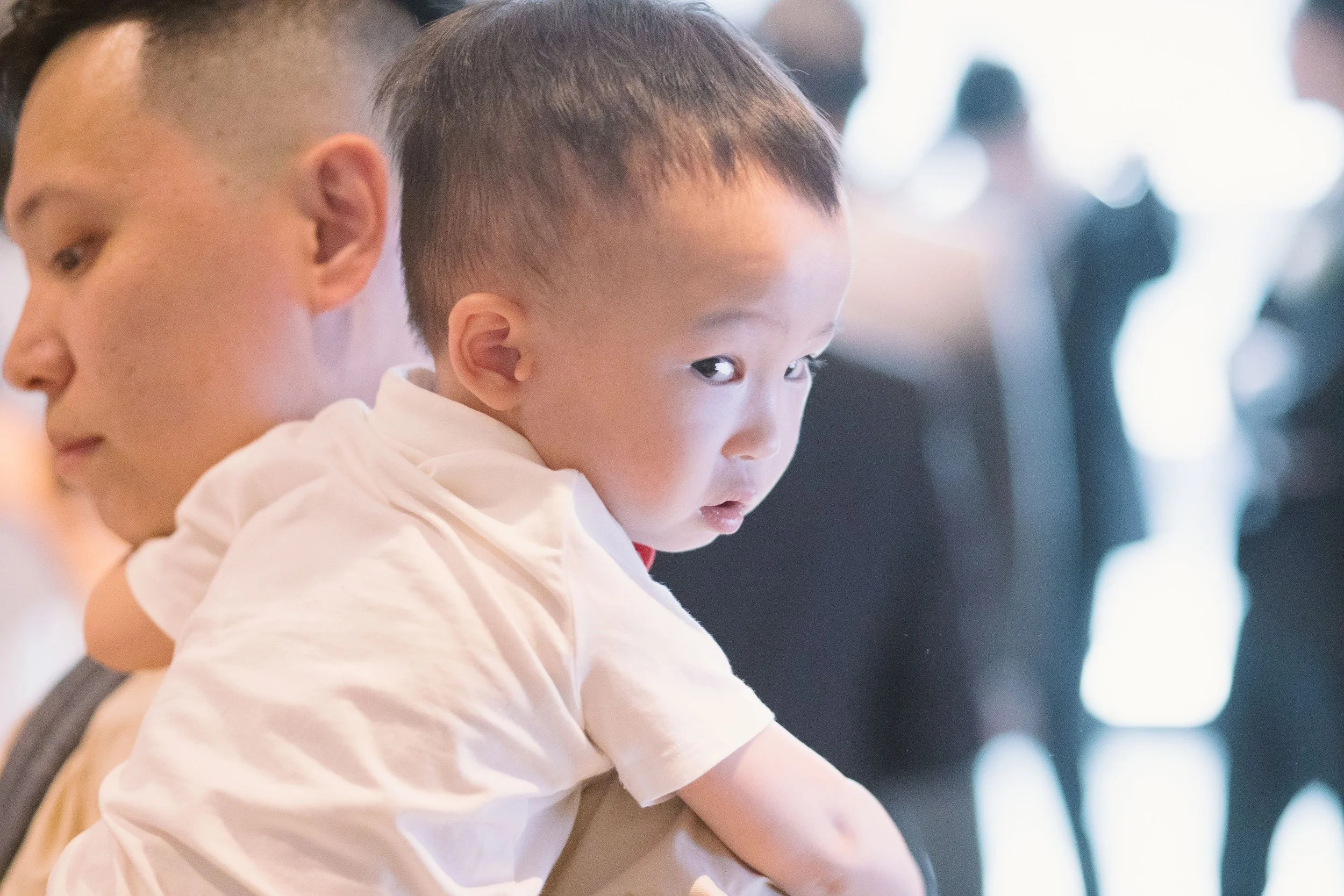 A young Asian boy with short black hair looking intently at something, sitting beside an older person with short dark hair, in a bright indoor setting with blurred background figures.