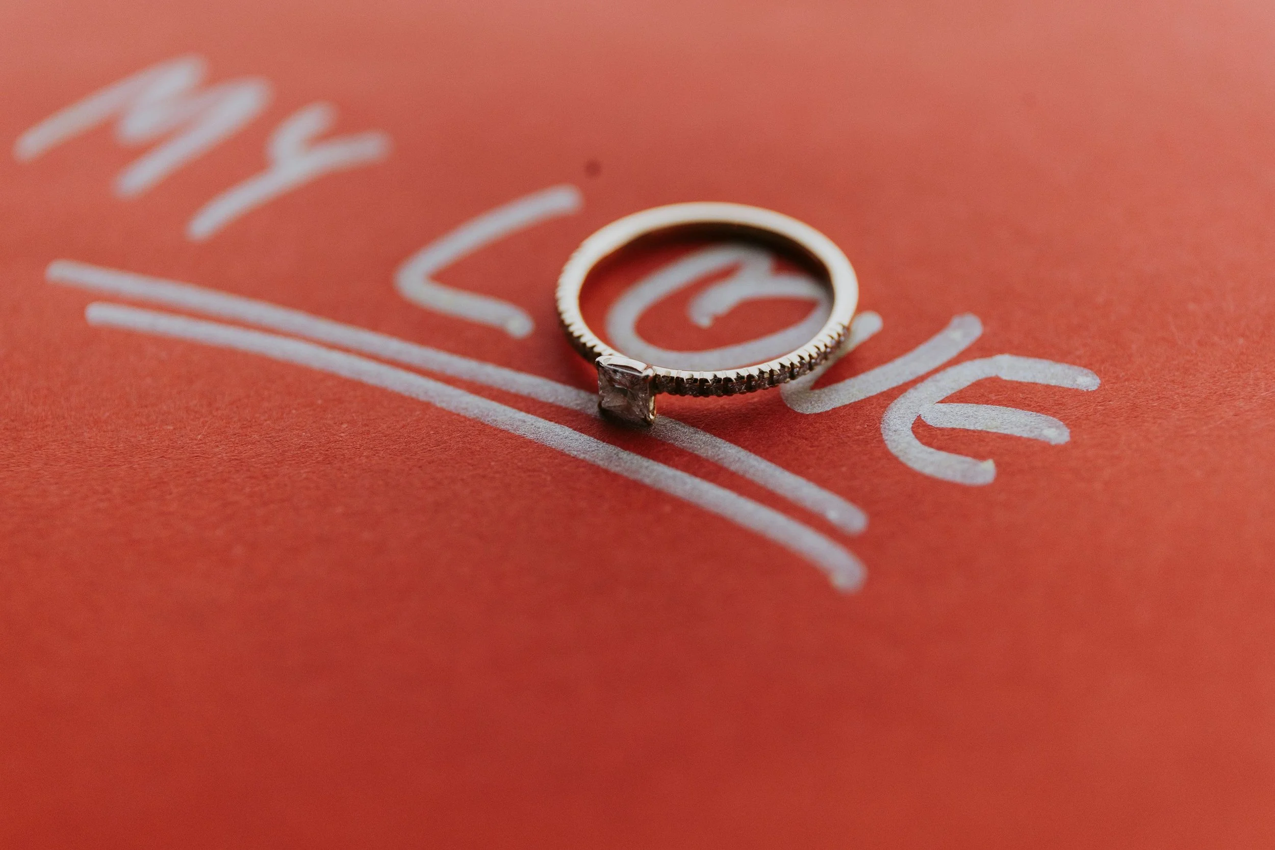 Close-up of a silver ring with a square-cut gemstone, resting on a red surface with white handwritten words, partly visible.
