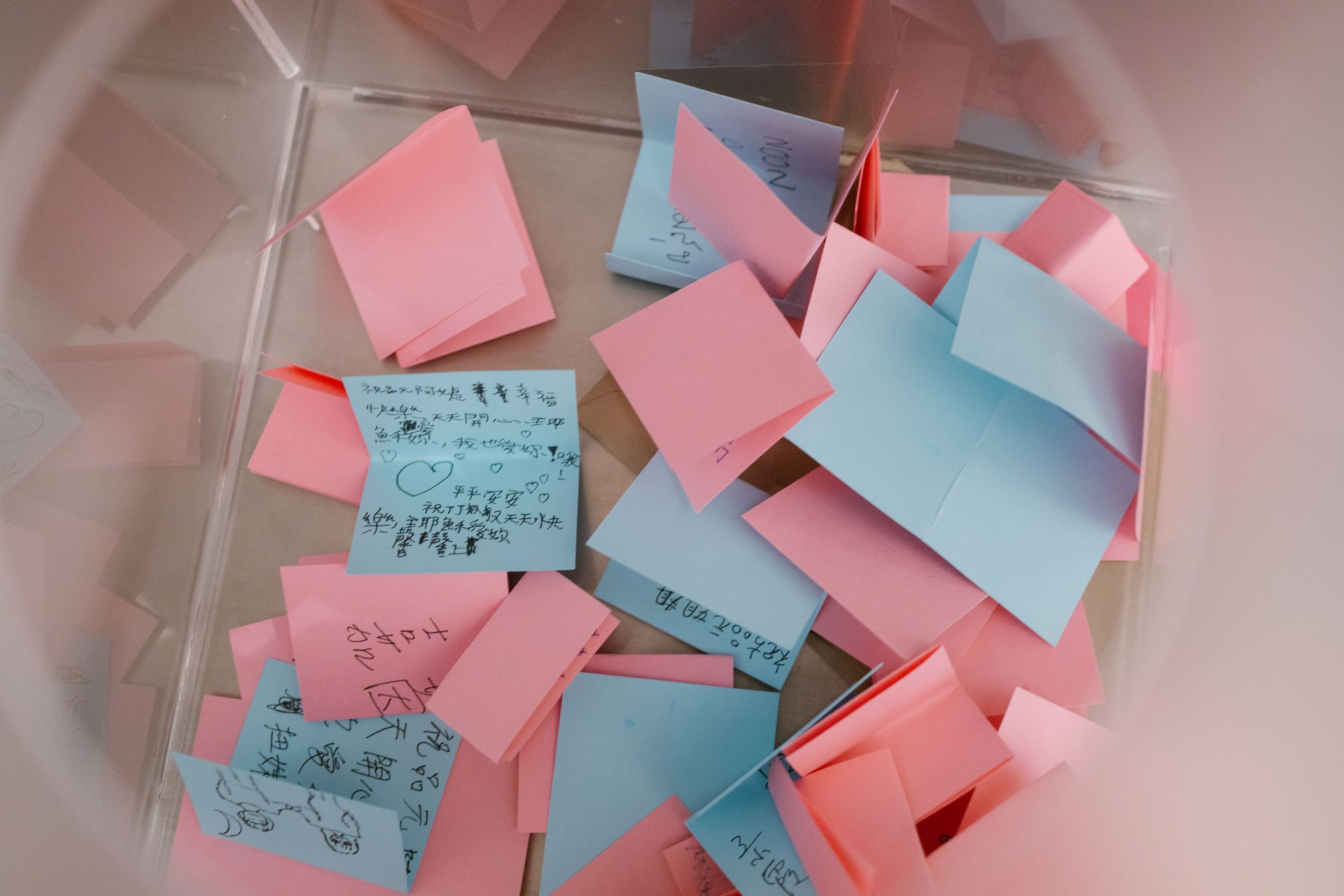 A transparent box filled with pink and blue sticky notes, some with handwritten messages and drawings.