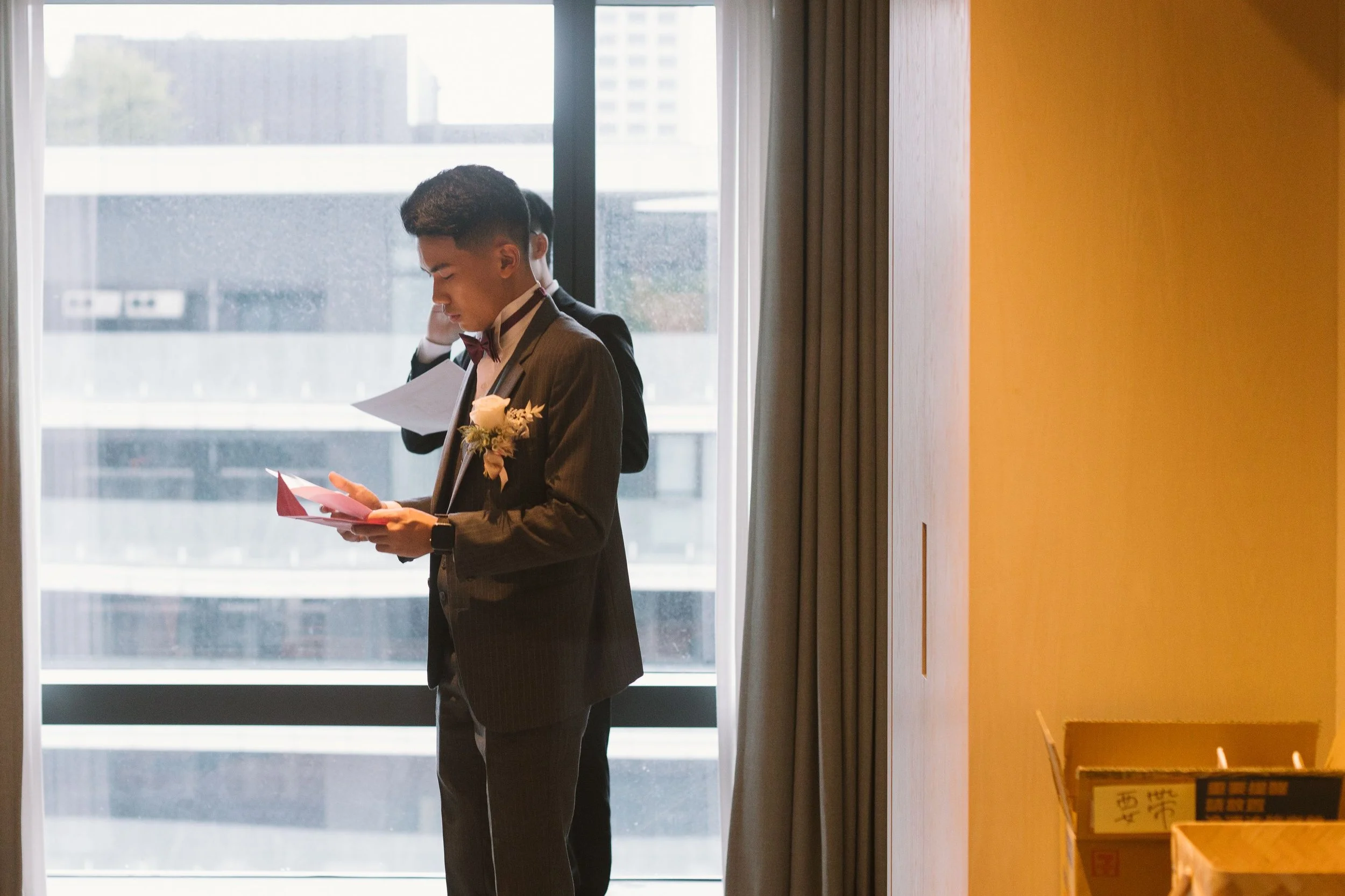 A man in a tuxedo with a boutonniere standing near a window, reading from a piece of paper, with another man in the background. The setting appears to be indoors, possibly during a formal event.