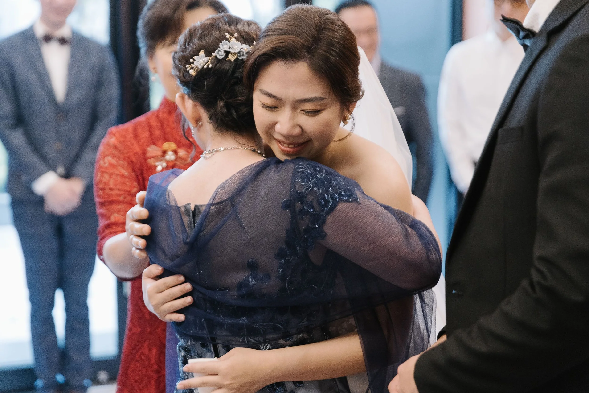 A woman in a dark blue dress with lace and embroidery is hugging another woman during a wedding or special event, with several people in formal attire in the background.