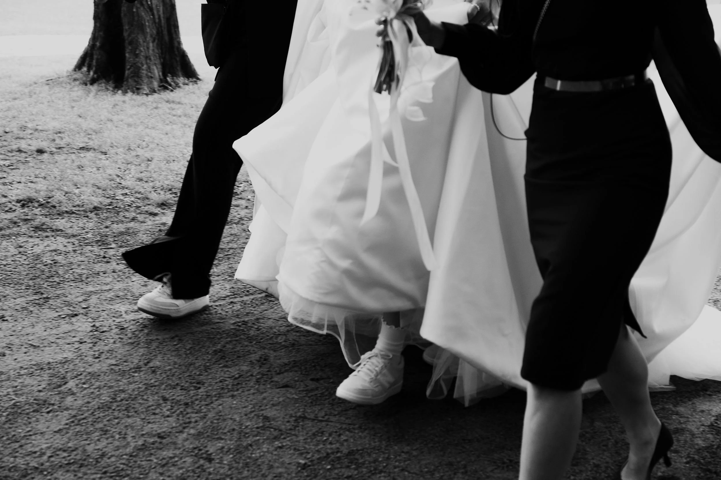 People wearing dresses and sneakers walking outdoors on a gravel path, with a tree in the background.