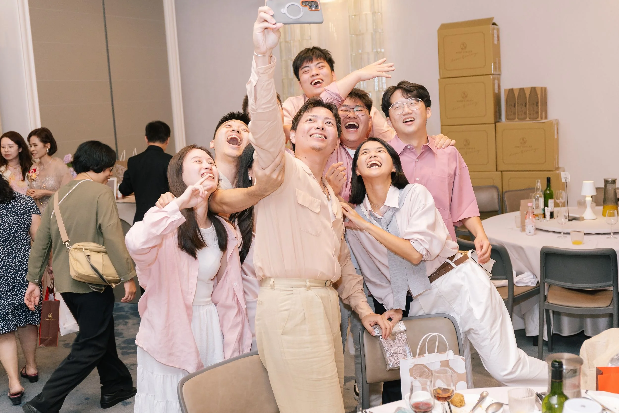 A group of people, mostly young adults, is taking a selfie together at a social gathering or party. They are smiling and laughing, with some making playful gestures. In the background, there are other guests, tables with food and drinks, and cardboar