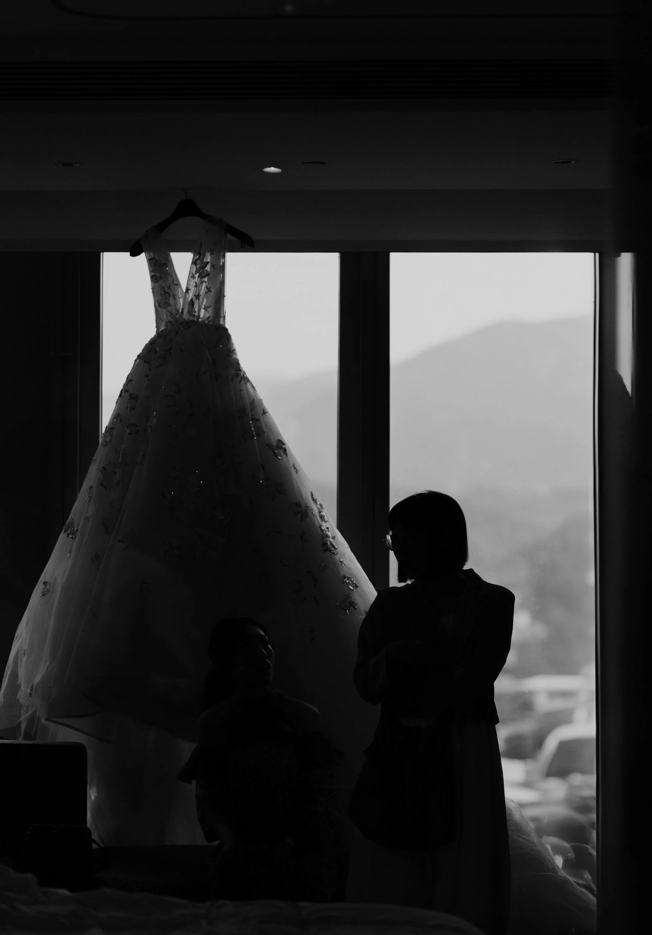 Silhouettes of children and adults observing a wedding dress hanging in front of a large window.