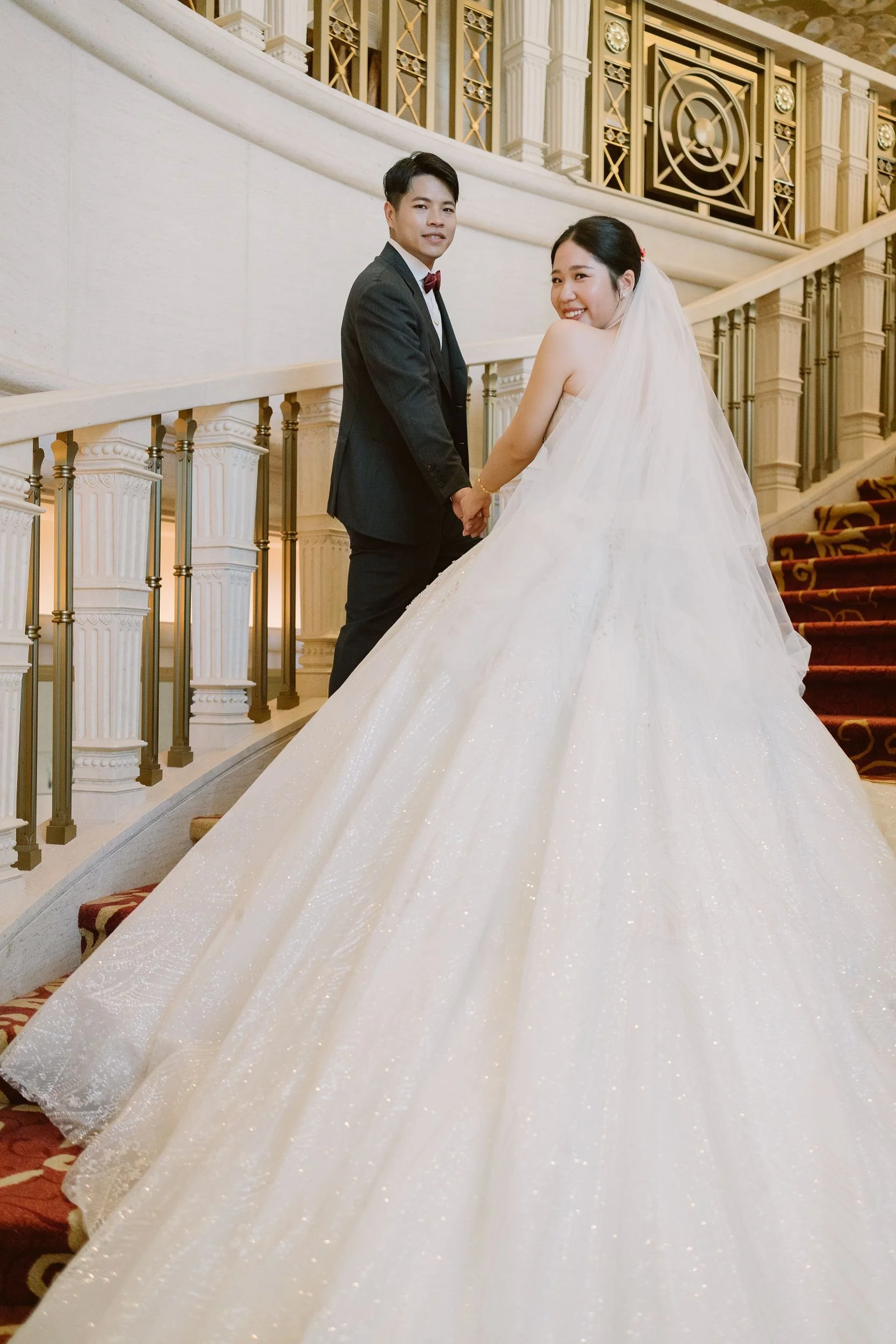 A bride and groom standing on a grand staircase inside a luxurious building, holding hands, with the bride smiling and wearing a strapless white wedding gown and veil, and the groom in a dark suit with a bow tie.