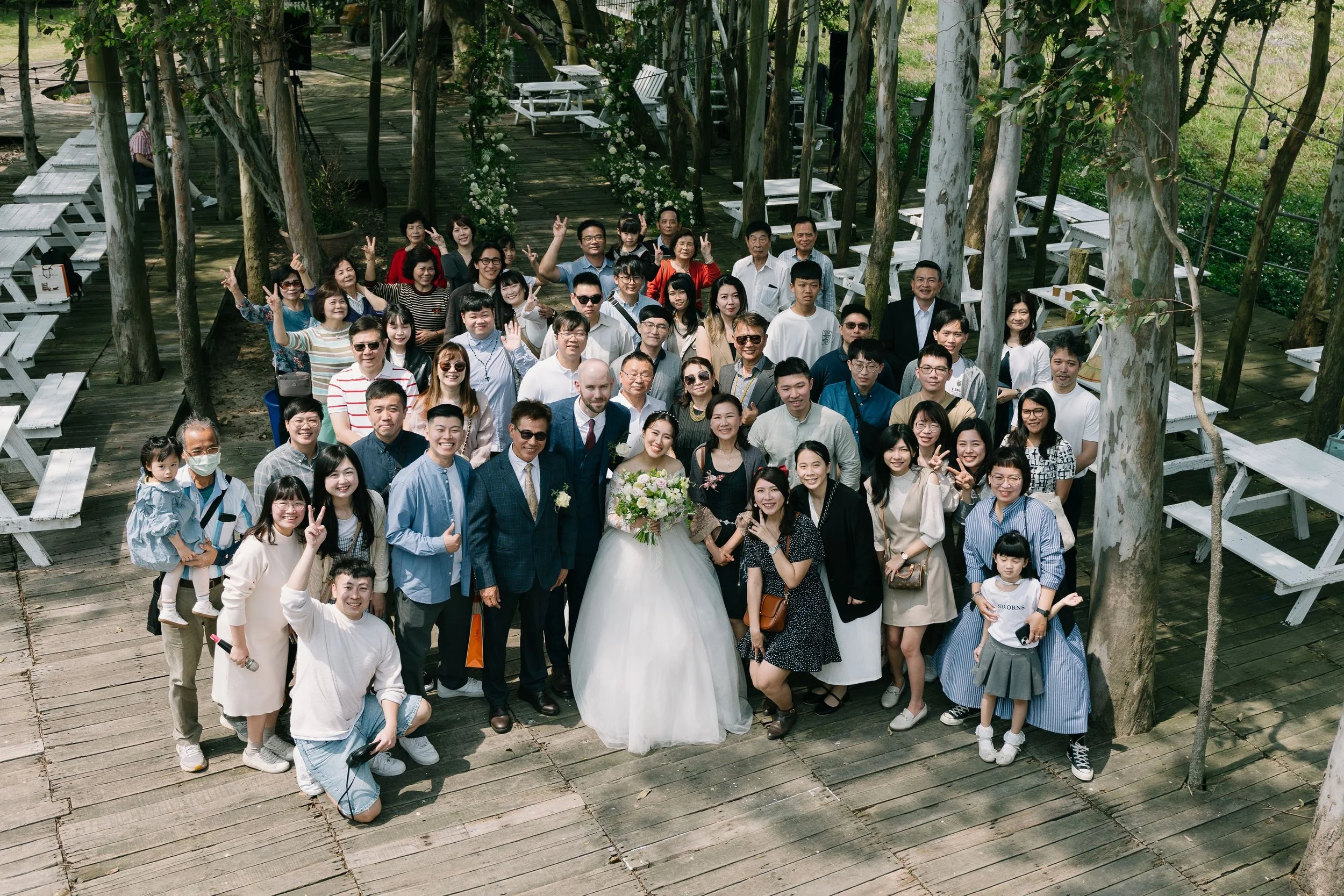Group of people gathering outdoors on wooden deck, posing for a photo, with some making peace signs, including a bride in a white wedding dress holding a bouquet, and a groom in a suit, surrounded by family and friends.