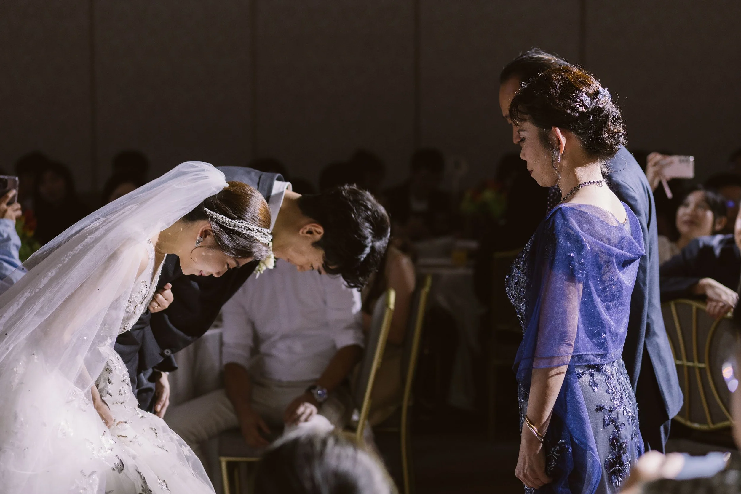 A bride and groom bowing during a wedding ceremony while guests look on in a dimly lit room.
