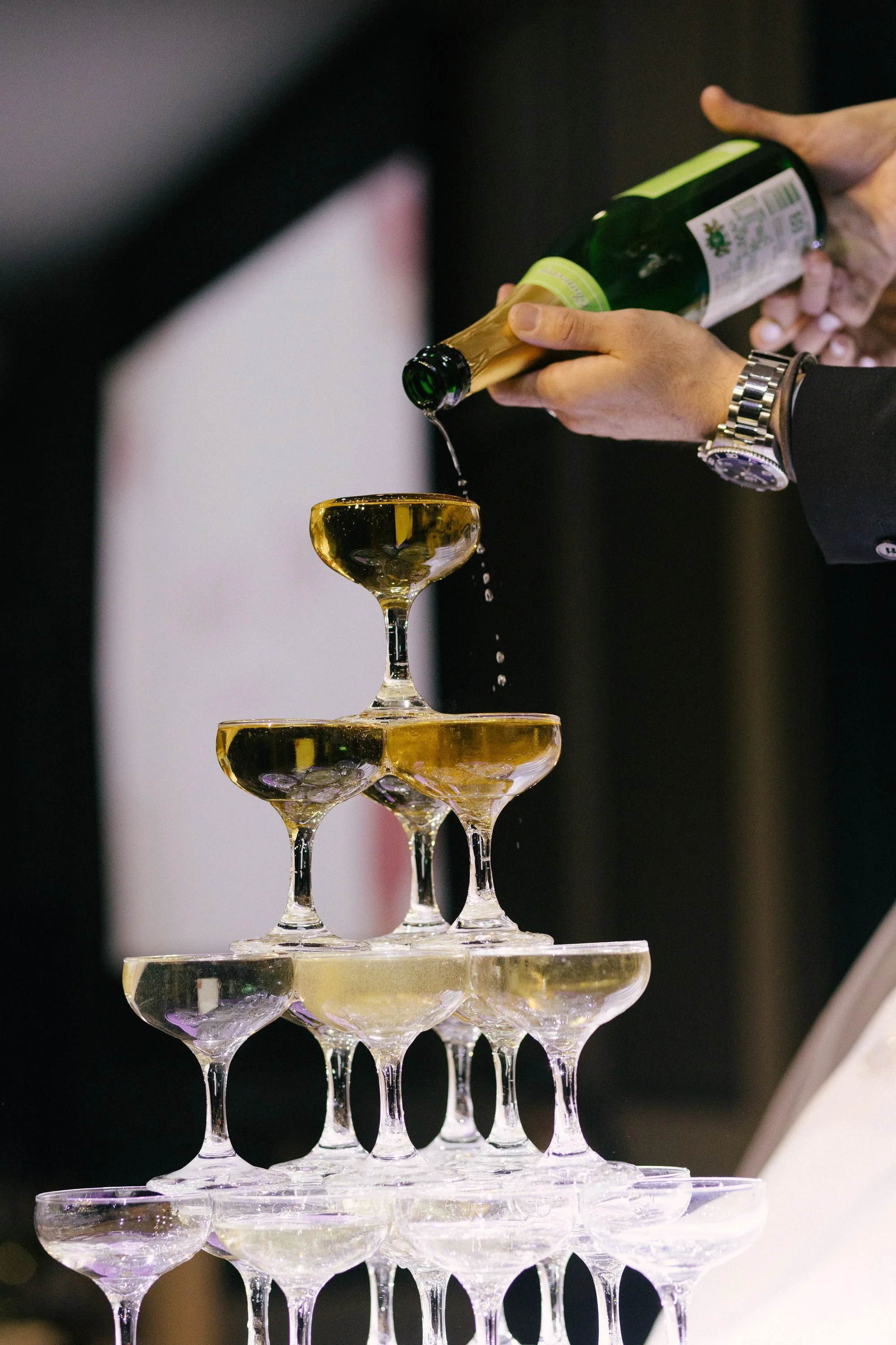 A champagne tower with multiple tiers of glasses being filled with champagne poured from a green bottle by a person wearing a black suit and silver watch.