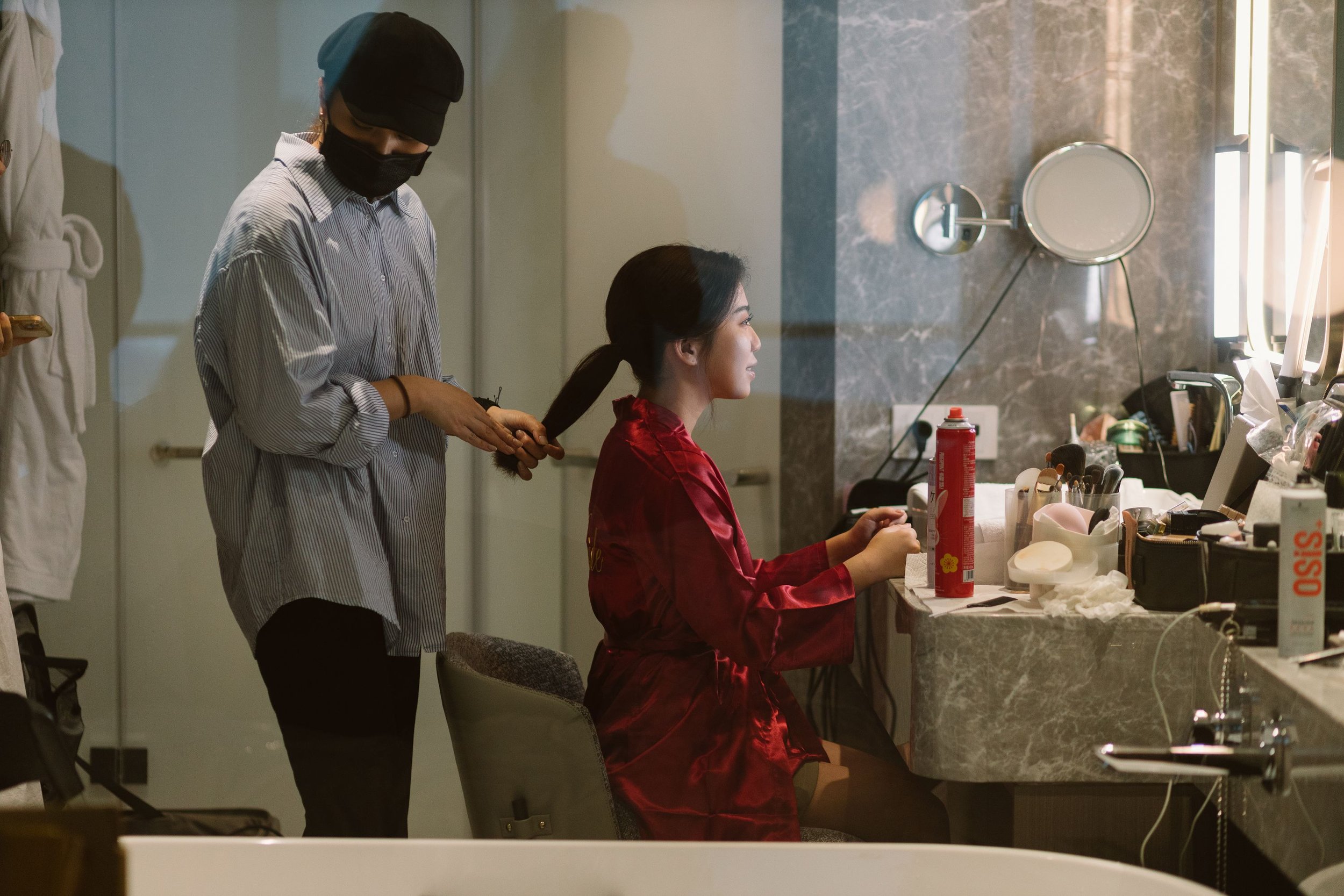 A woman sitting at a vanity mirror having her hair styled by a hairstylist, both wearing masks. The woman has a smile and is wearing a red satin robe. The vanity table has various makeup and hair styling tools.