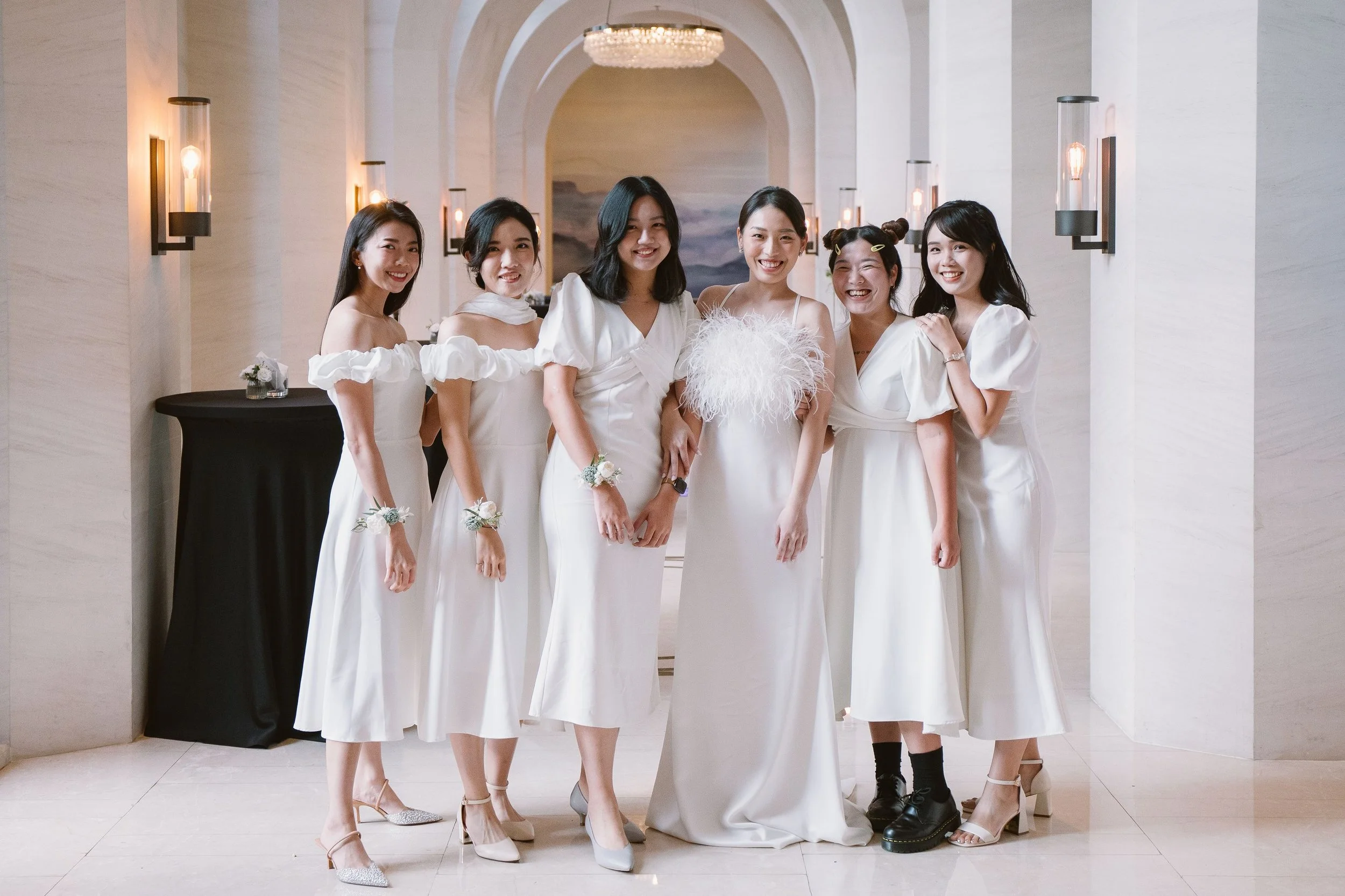 A group of six women dressed in white, standing in a decorated indoor space with arched ceilings and wall sconces, smiling at the camera.