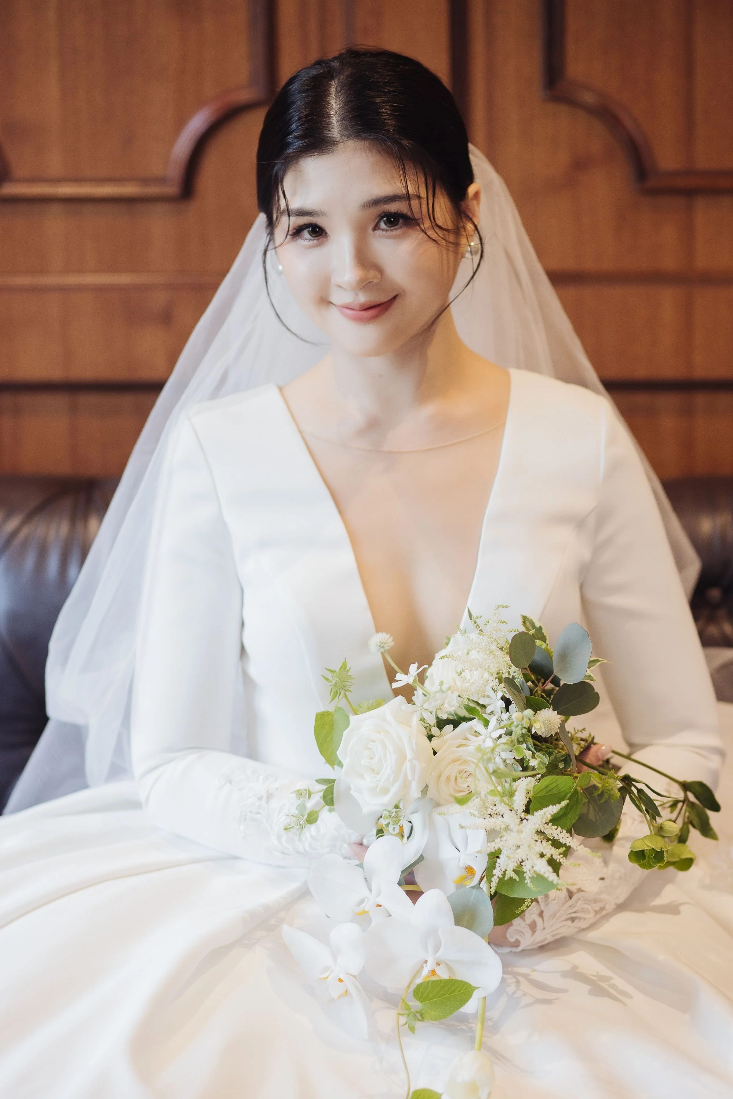 A young woman in a wedding dress sitting and holding a bouquet of white flowers, with a wooden background.