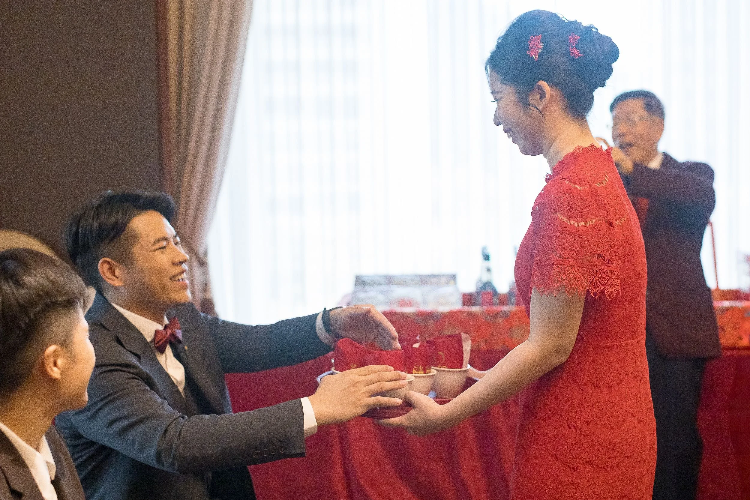 A woman in a red lace dress is smiling and shaking hands with a man in a gray suit and bow tie, who is seated at a table. The shirt of someone leaned over is also visible in the foreground. In the background, an older woman in glasses stands and look