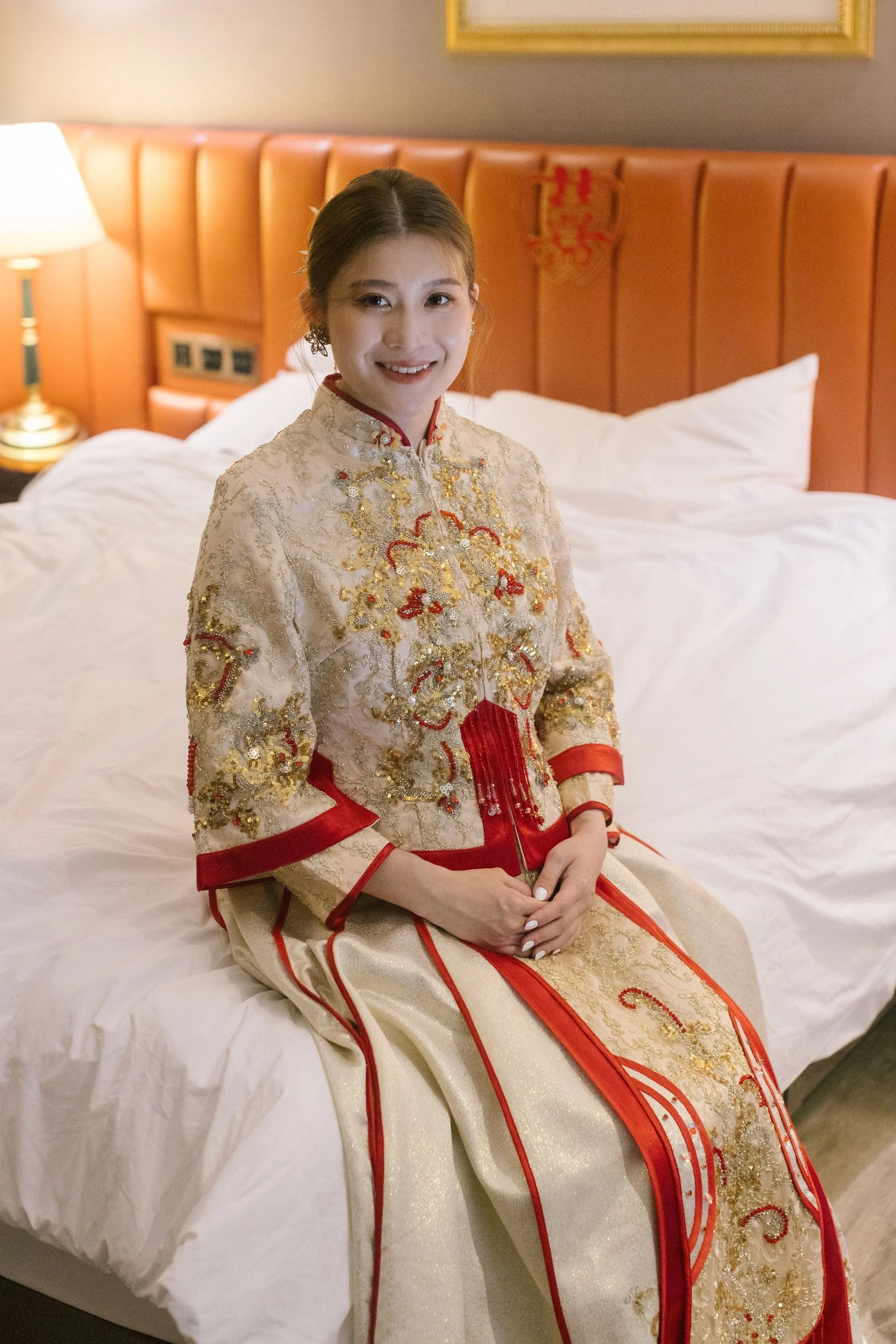 A woman dressed in a traditional Chinese wedding dress sitting on a bed in a hotel room, smiling at the camera.
