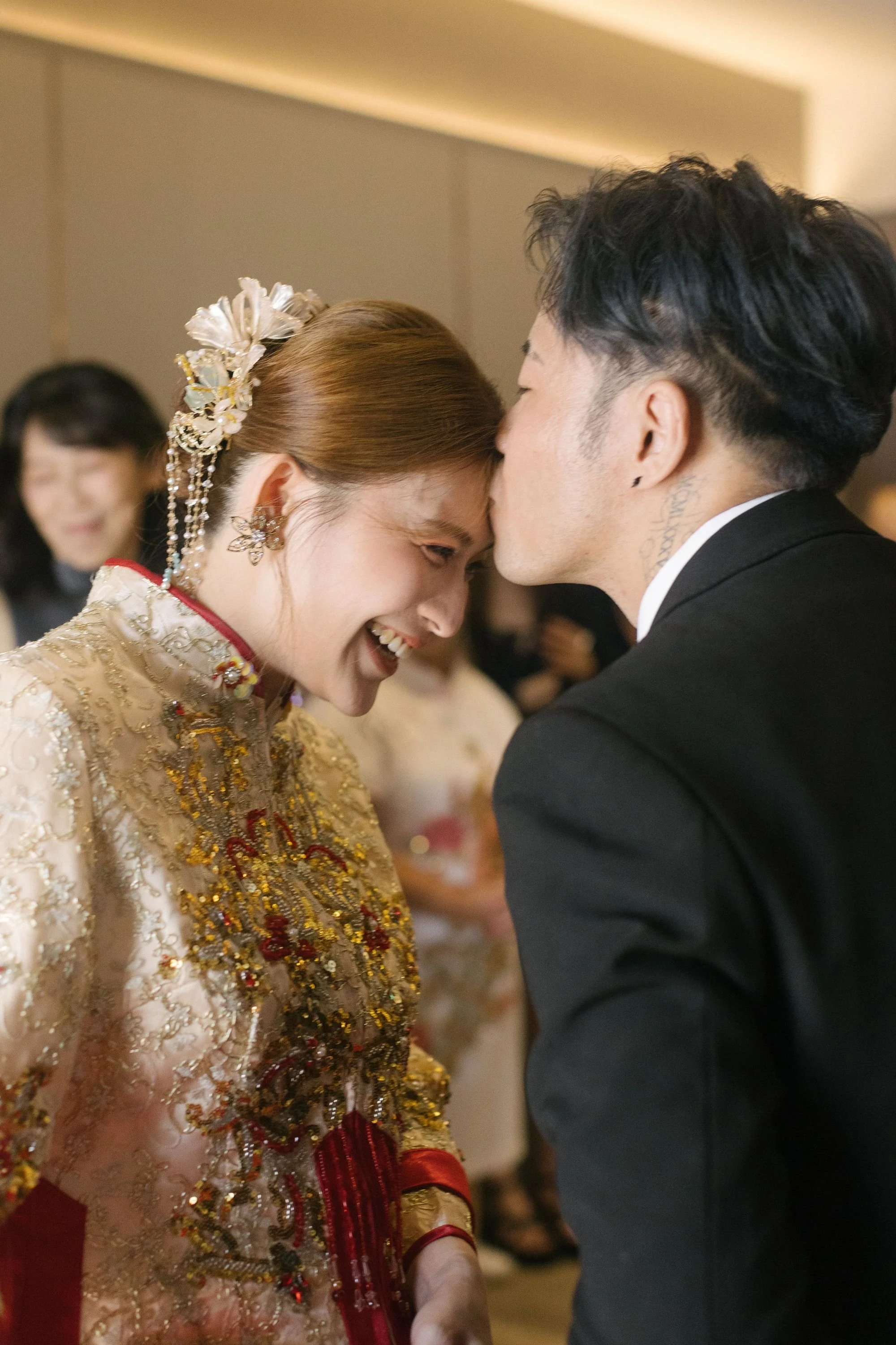 A woman and a man are touching foreheads and smiling at each other at a formal event. The woman is wearing traditional attire with intricate embroidery and jewelry, and the man is in a black suit.