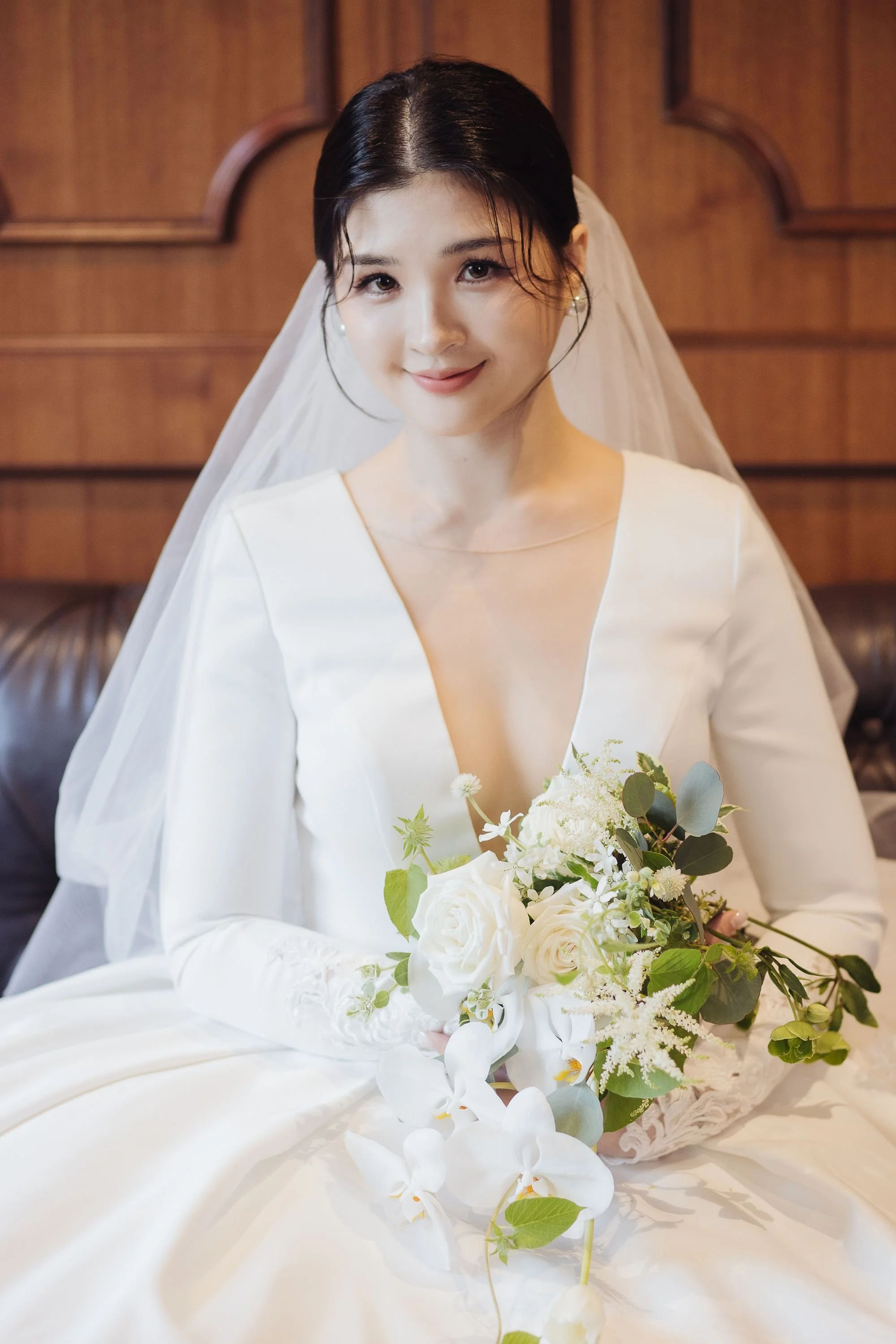 A woman in a wedding dress holding a bouquet of white flowers, sitting against a wooden background.