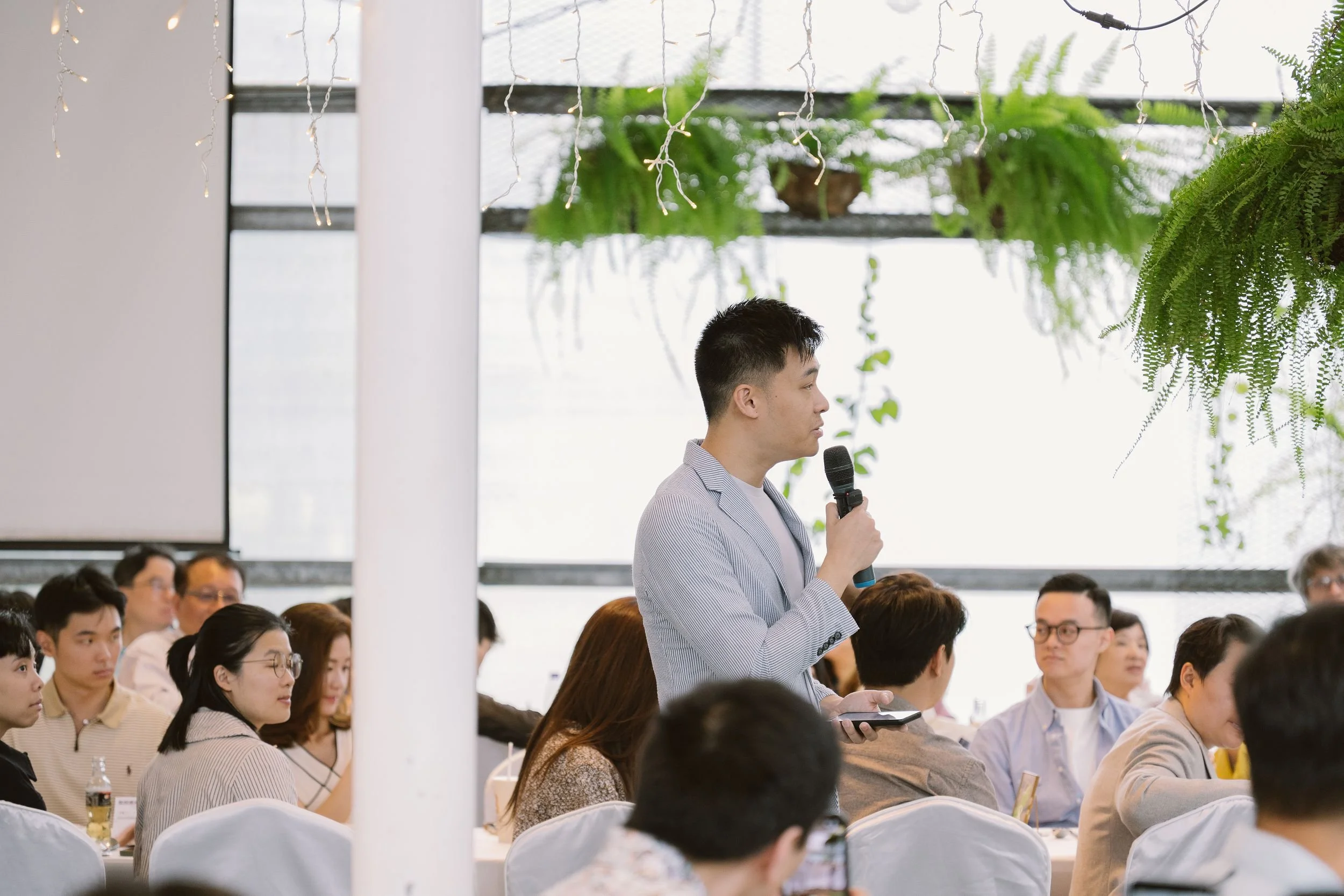 A man standing and speaking into a microphone during a conference or seminar, with audience members seated and listening attentively in a bright room decorated with hanging string lights and lush green plants.