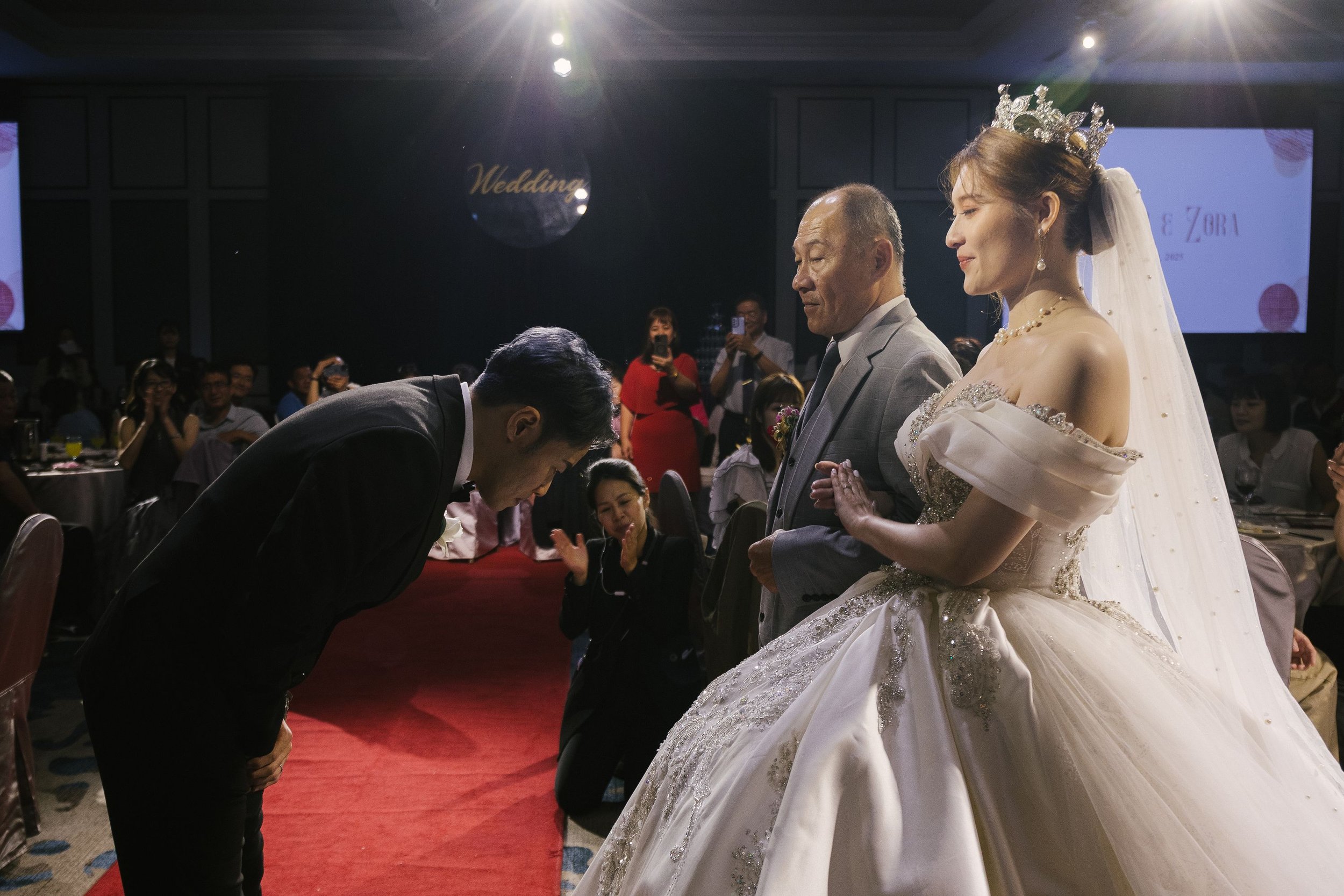 A bride in a wedding dress and a tiara with a veil, holding hands with an older man in a suit, during a wedding ceremony. A man is bowing deeply in front of them, and several guests are visible in the background, some taking photos, at a decorated we