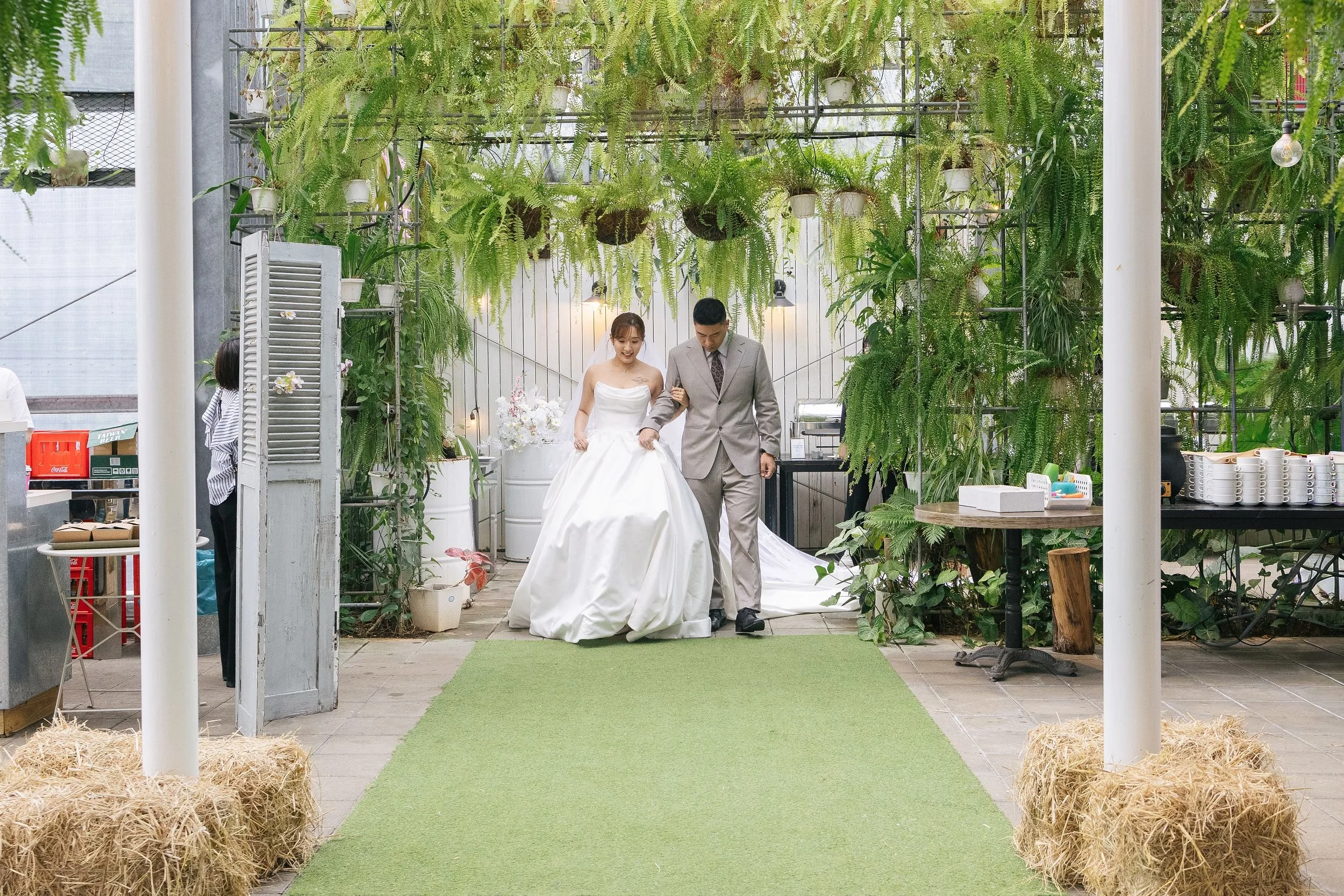 A bride and groom walking down an aisle decorated with greenery and plants in a greenhouse or plant shop.