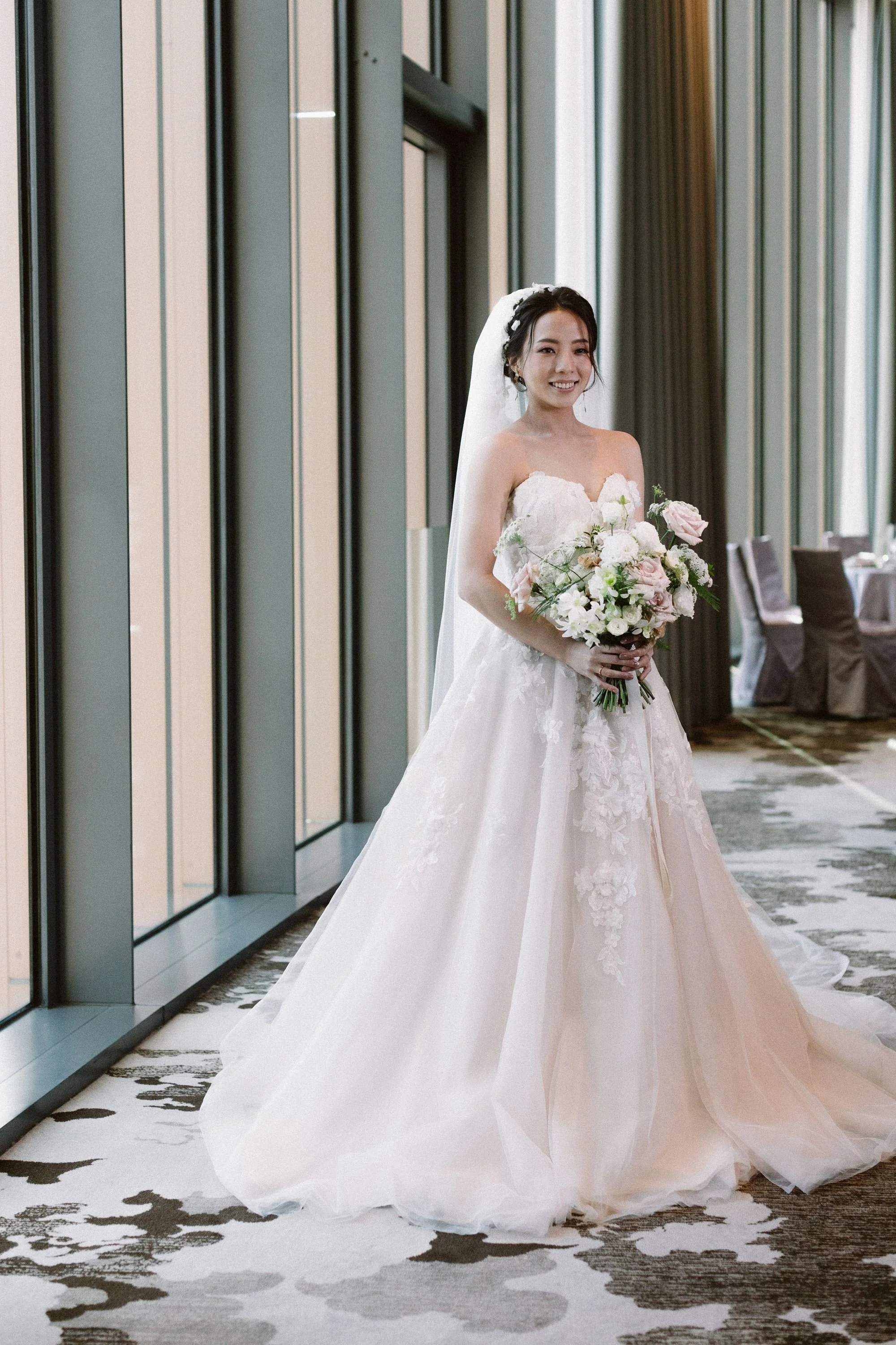 A bride in a white wedding gown holding a bouquet of pink and white flowers, standing indoors near glass walls, smiling.