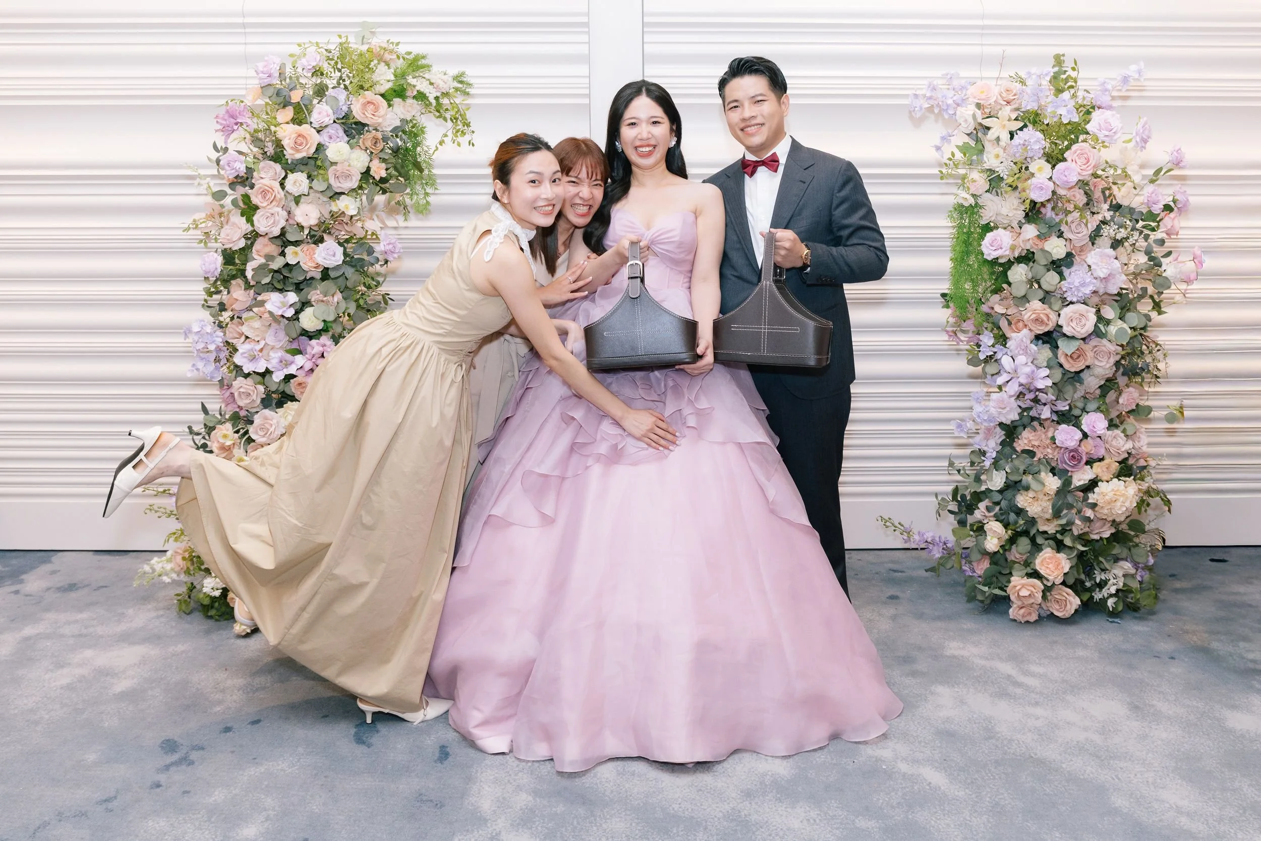 Group of five happy people celebrating at a wedding, standing in front of floral arrangements with pastel-colored roses and hydrangeas, smiling and posing for a photo.