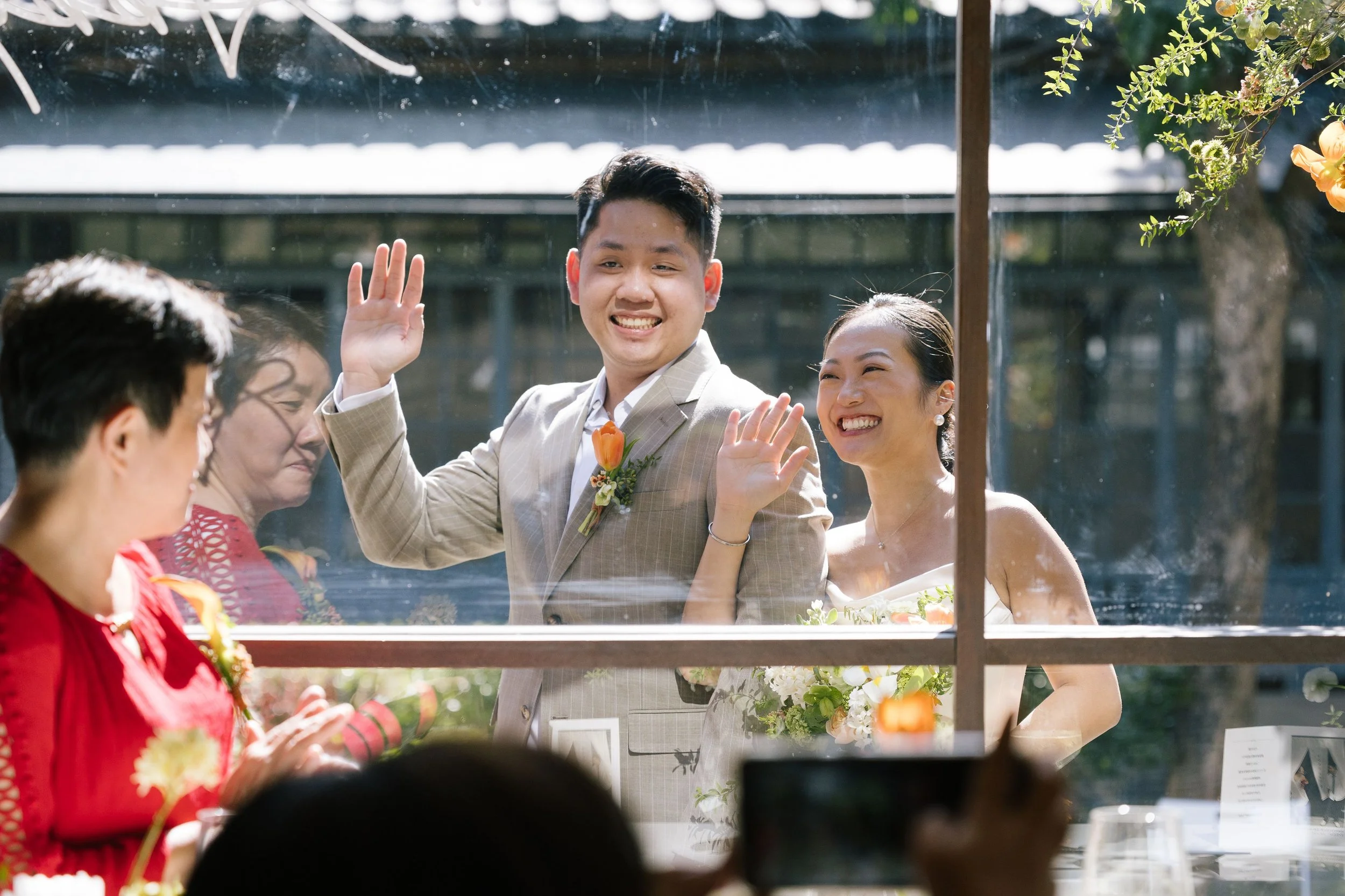 A group of people enjoying a celebration, with a man in a beige suit waving through a glass window, and women dressed in festive attire, smiling and holding flowers, outdoors on a sunny day.