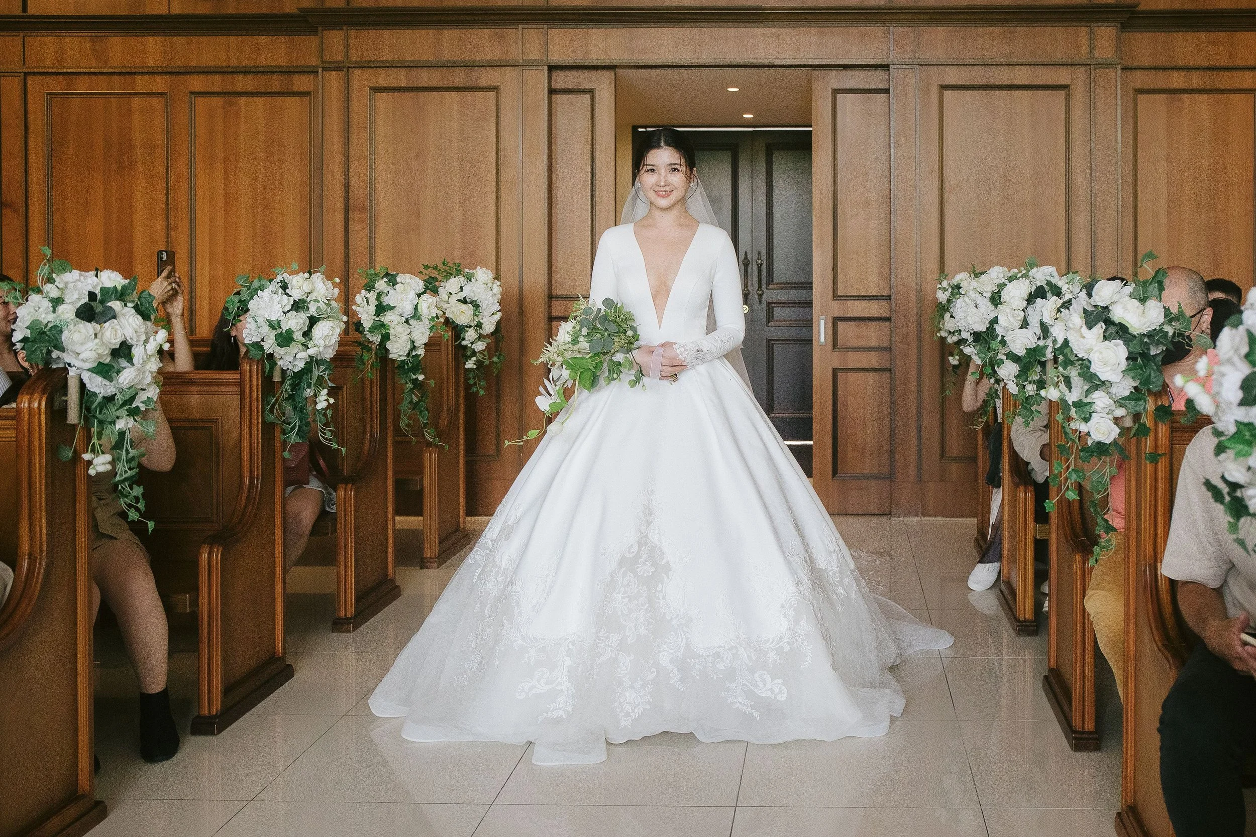 A bride in a white wedding gown holding a bouquet of greenery and flowers, standing in a wooden-paneled room decorated for a wedding ceremony, with guests seated on either side.