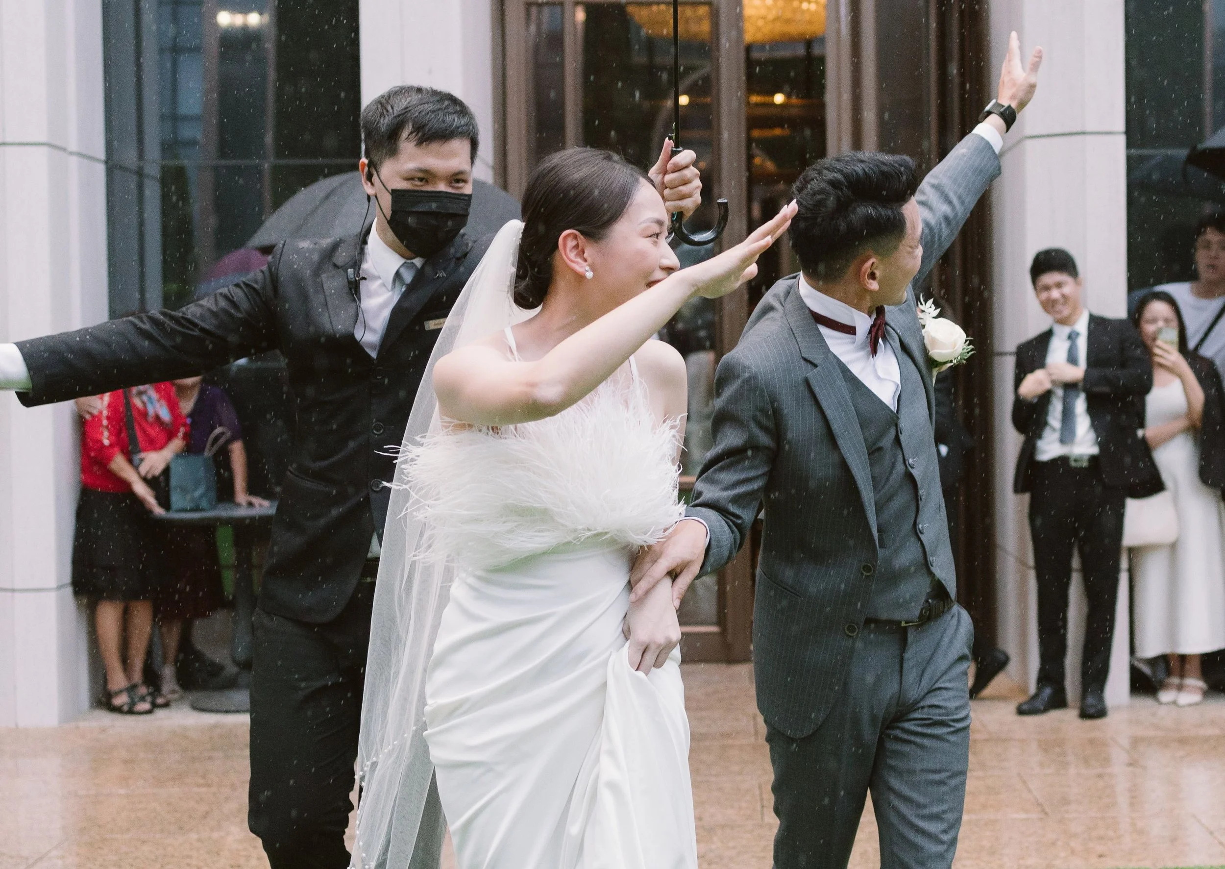 A bride in a white wedding dress with a fluffy feather detail on the side, smiling and saluting, is walking outdoors. She is holding hands with a groom in a gray suit with a boutonniere. A man in a suit and mask is holding an umbrella above them. In 