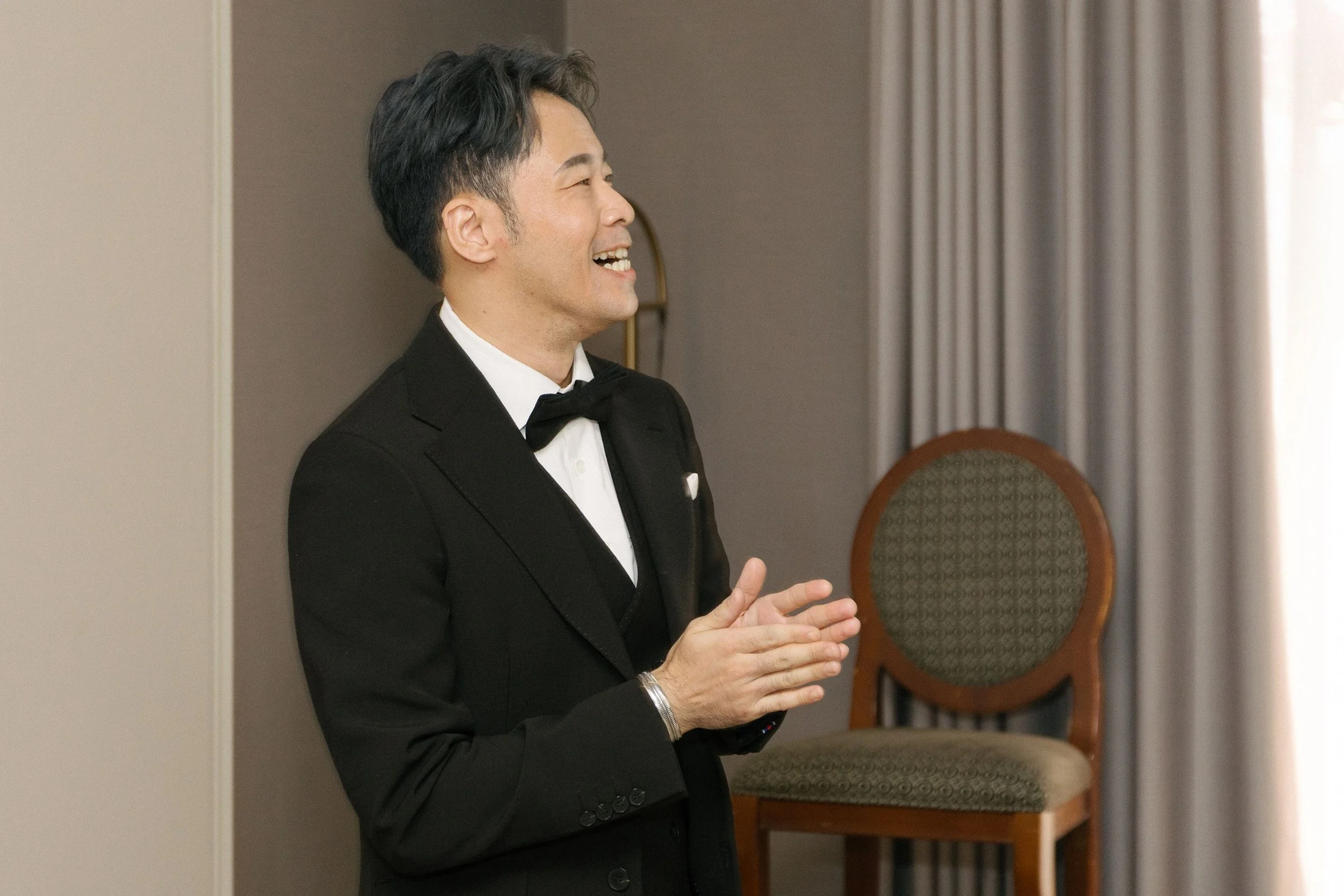 A man in a black tuxedo with a bowtie, smiling and clapping, standing indoors near chairs and curtains.