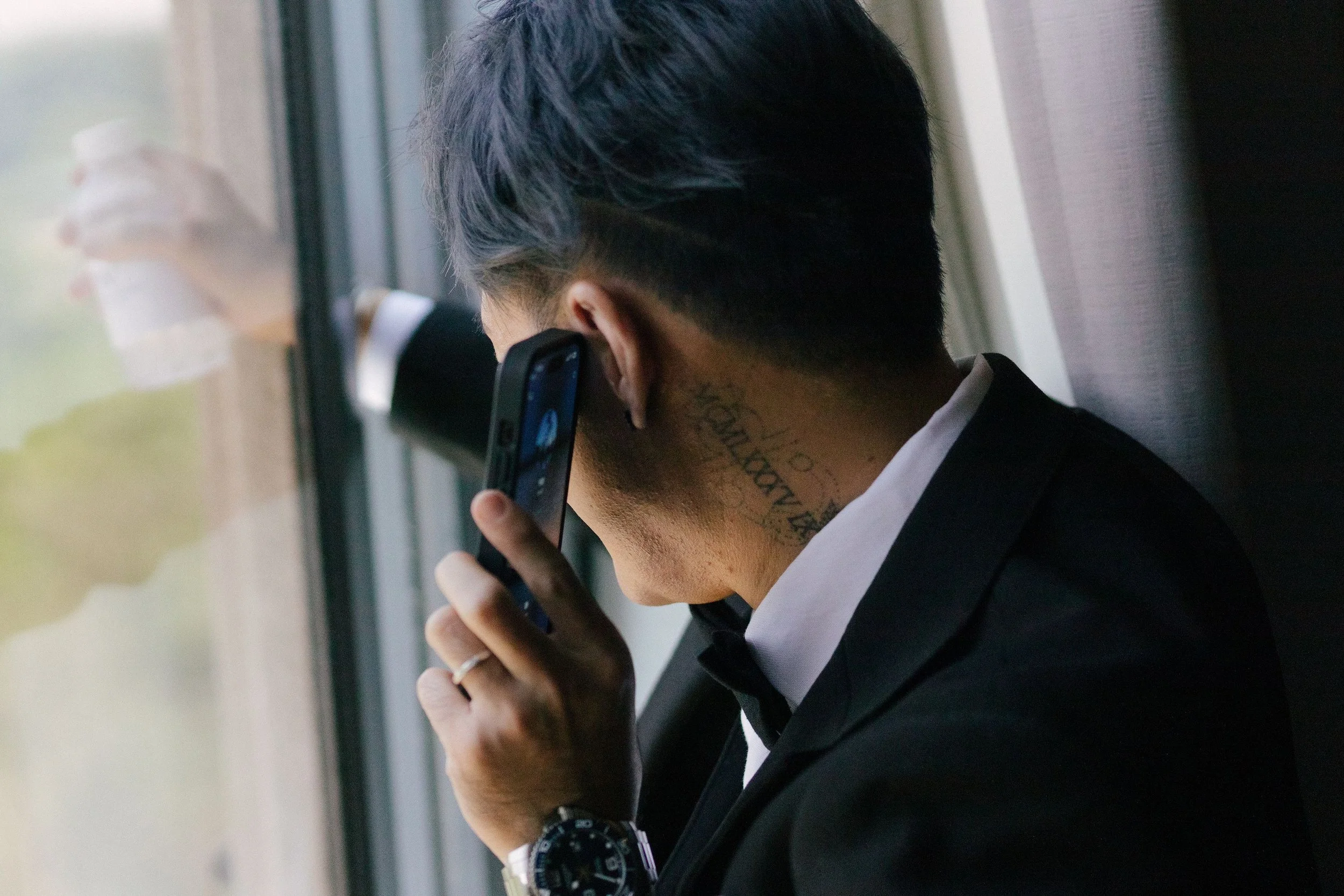 Person with dark hair, wearing a black suit, talking on a cellphone near a window.