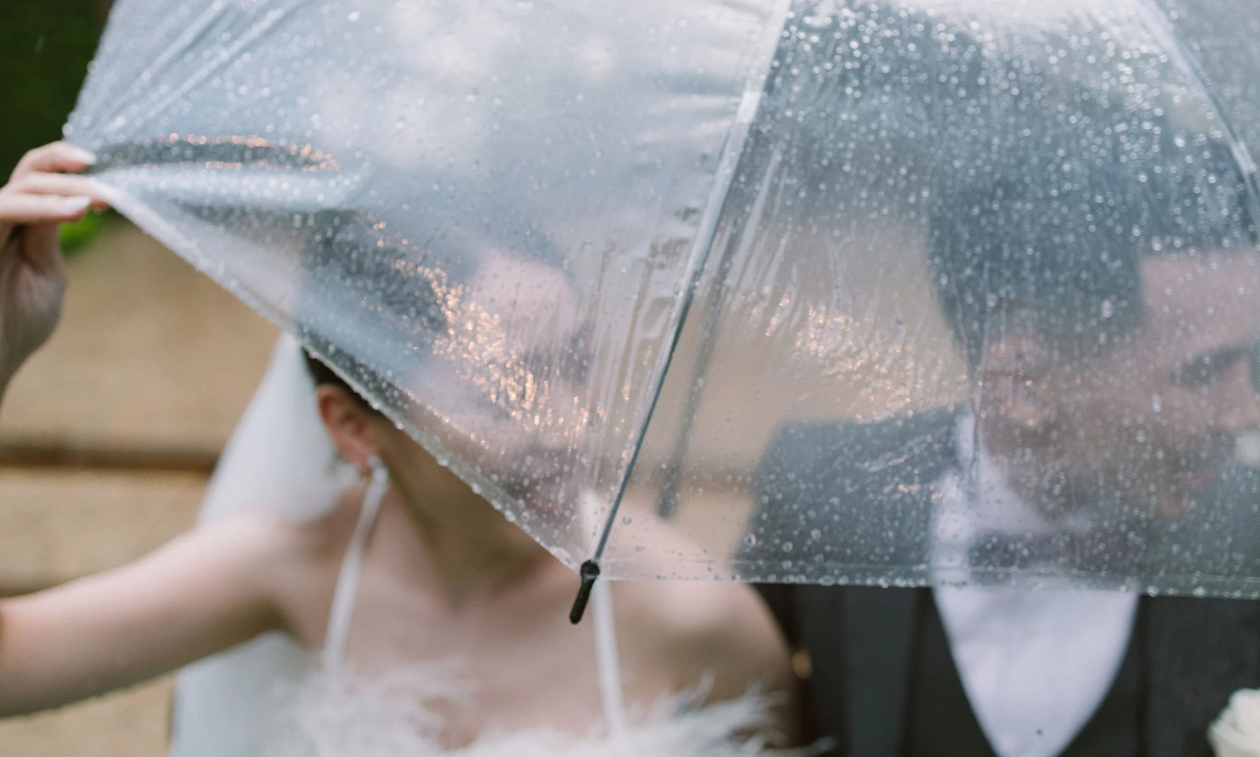 A person holding a clear umbrella with raindrops on it, partially covering their face, outdoors during rainy weather.