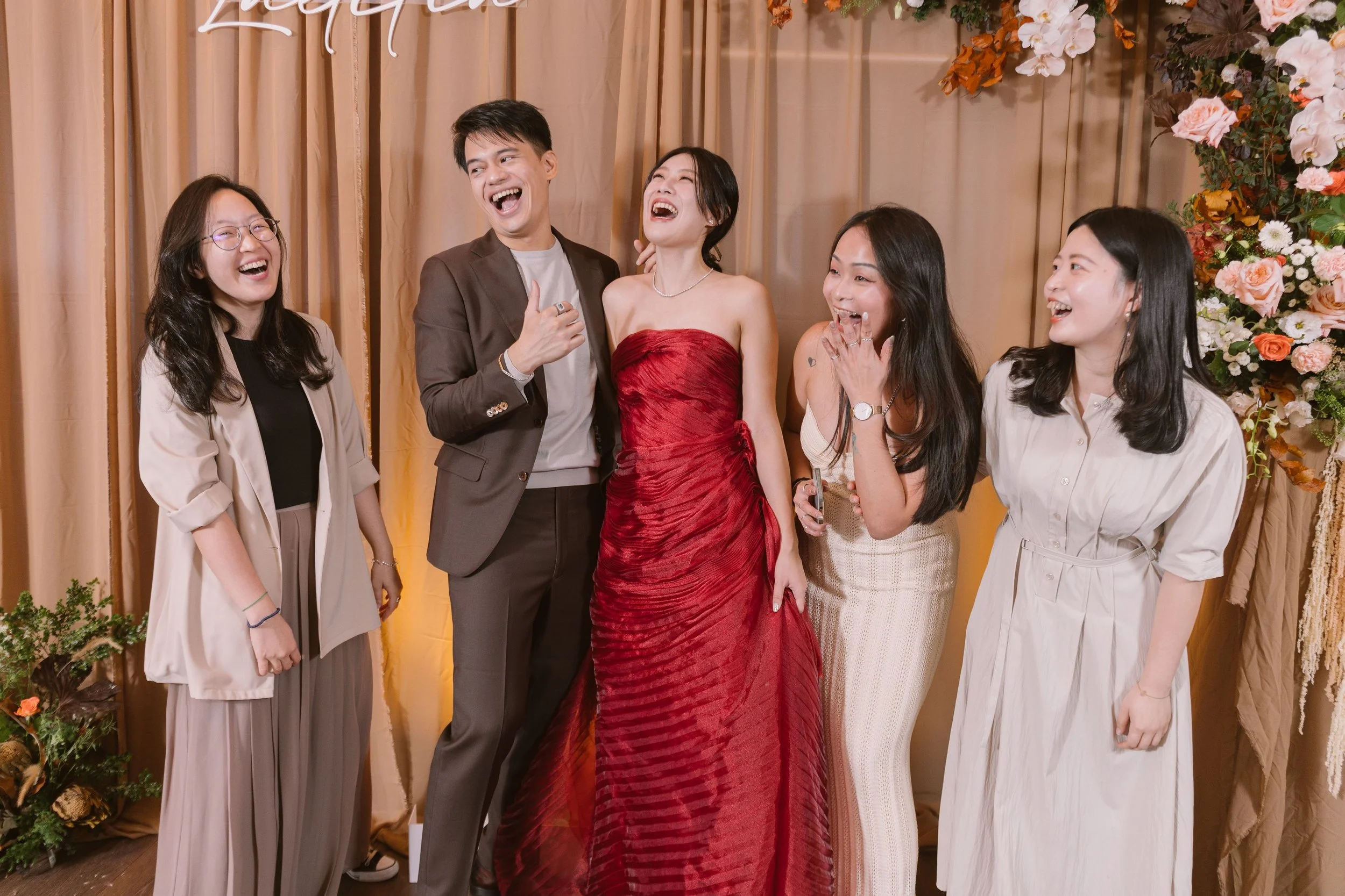 Group of five people, three women and two men, laughing and enjoying themselves at an event with a flower backdrop.