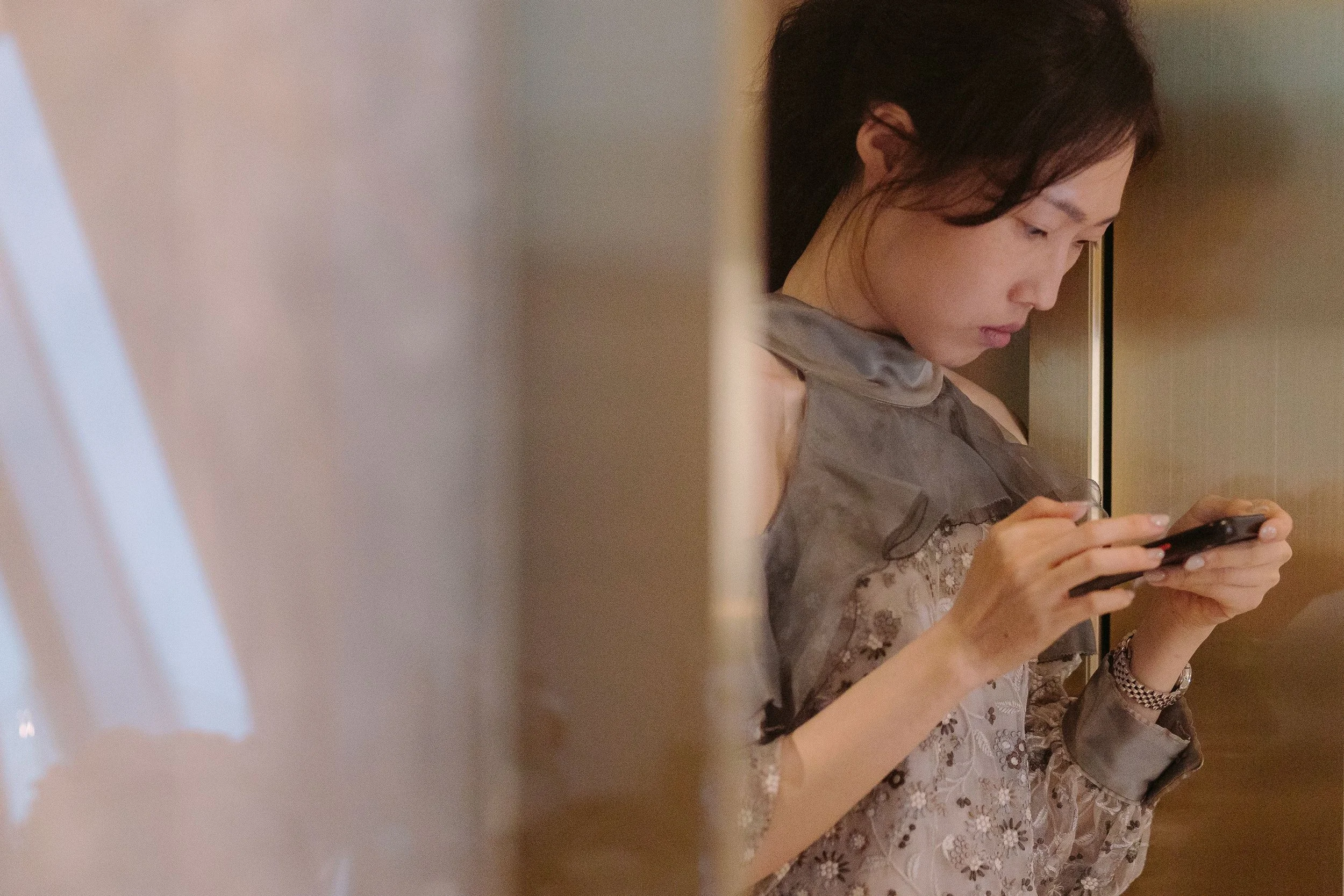 A woman with dark hair looking down at her phone, dressed in a floral lace top, sitting at a table, with a warm indoor lighting background.