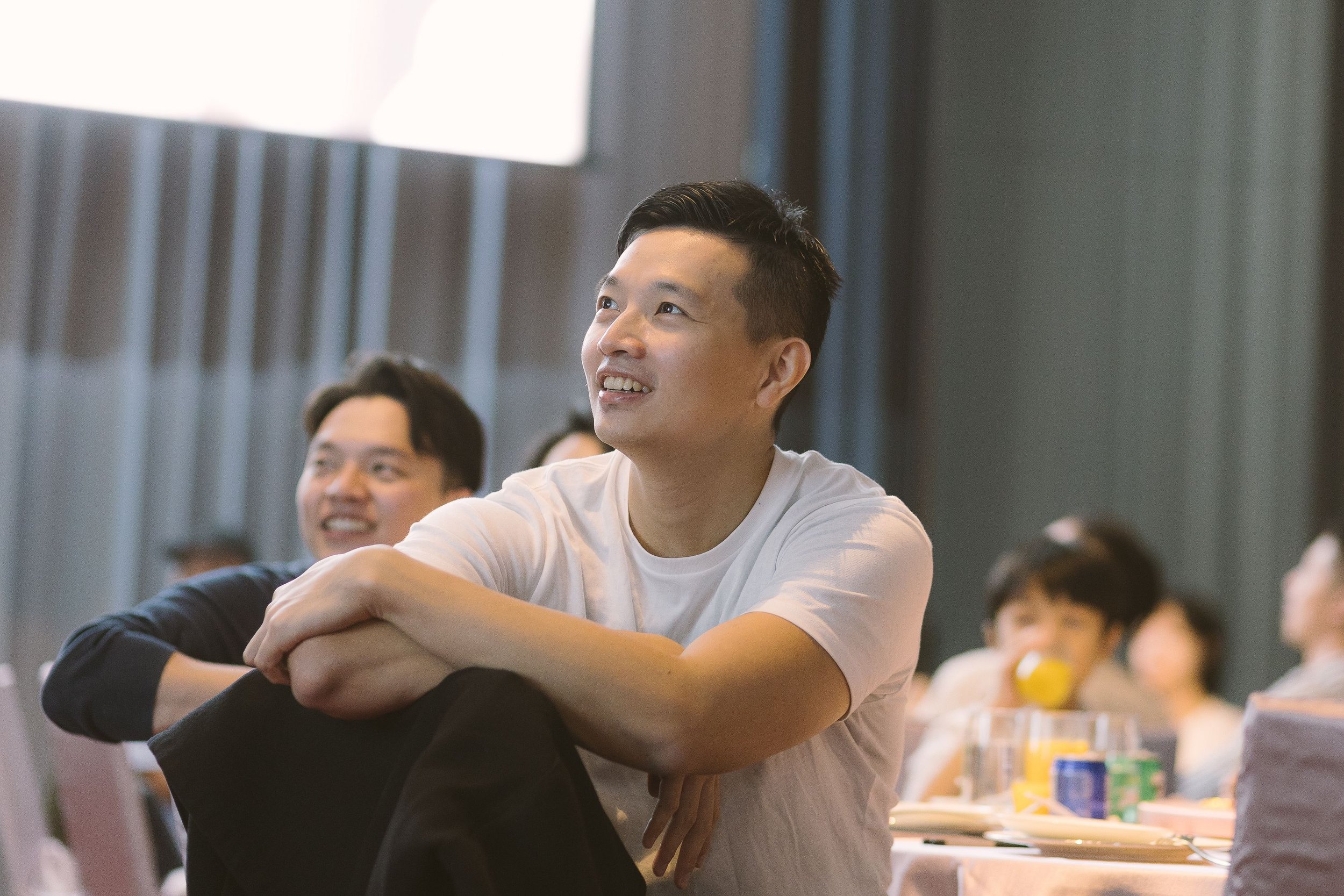 Young man in white T-shirt smiling, sitting with folded arms, at a table during a gathering or seminar, with others in the background.