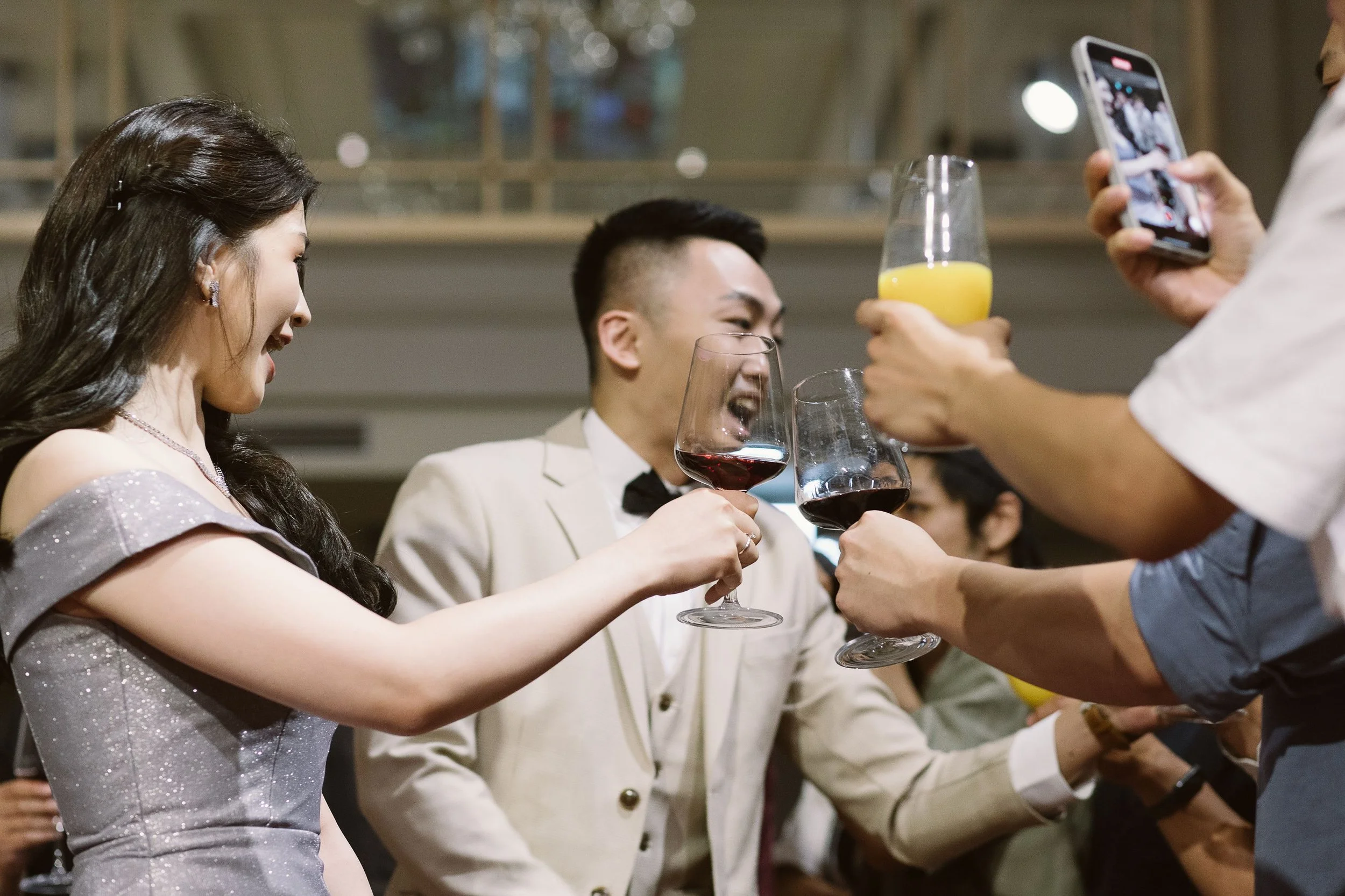 A group of people at a social gathering or party, raising glasses with drinks such as wine, juice, and water, clinking them together in a celebration. The individuals are dressed in formal and semi-formal attire, with one woman wearing a sparkly dres
