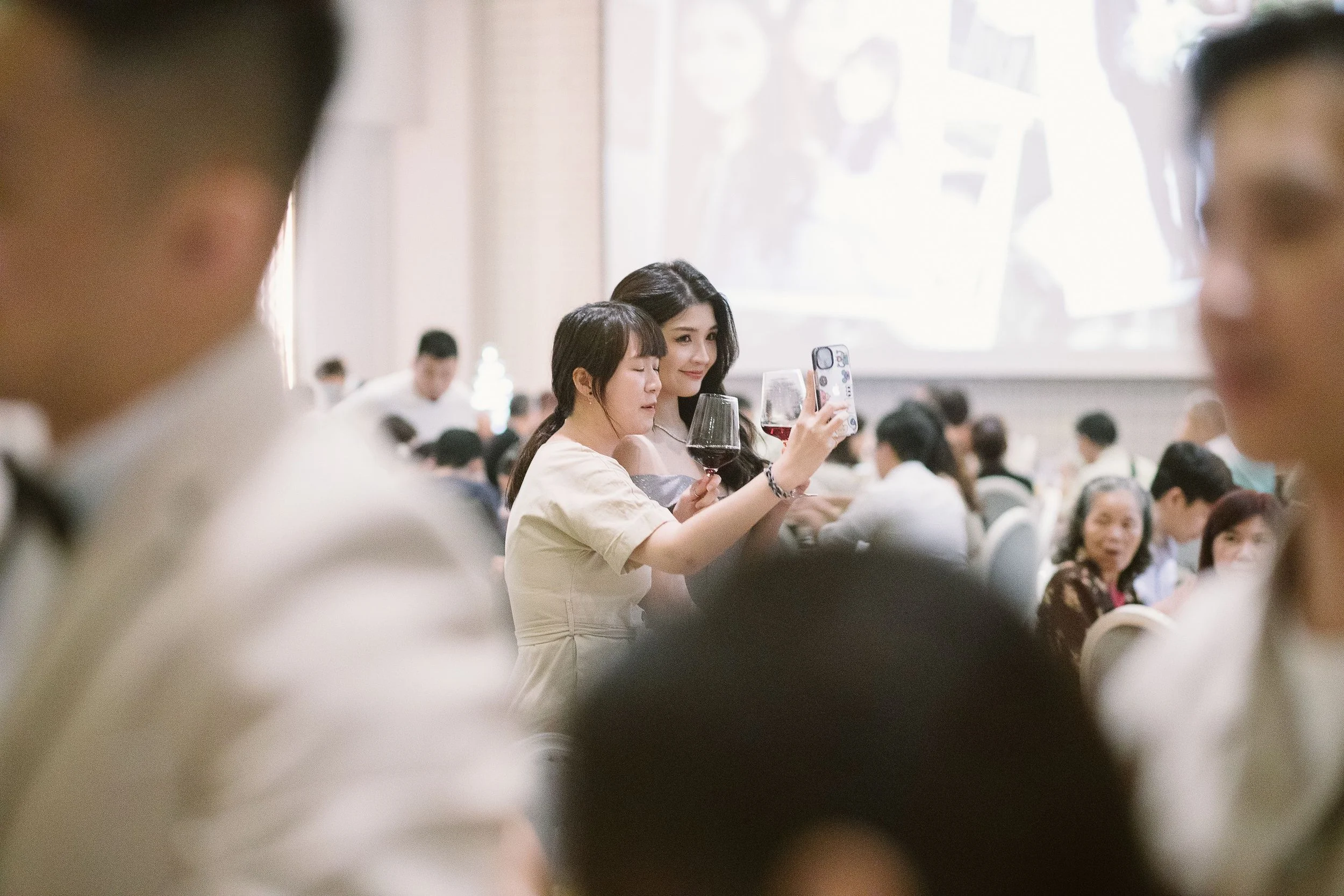 Two women taking a selfie at a crowded event with many people seated, some holding wine glasses, in a well-lit indoor setting.