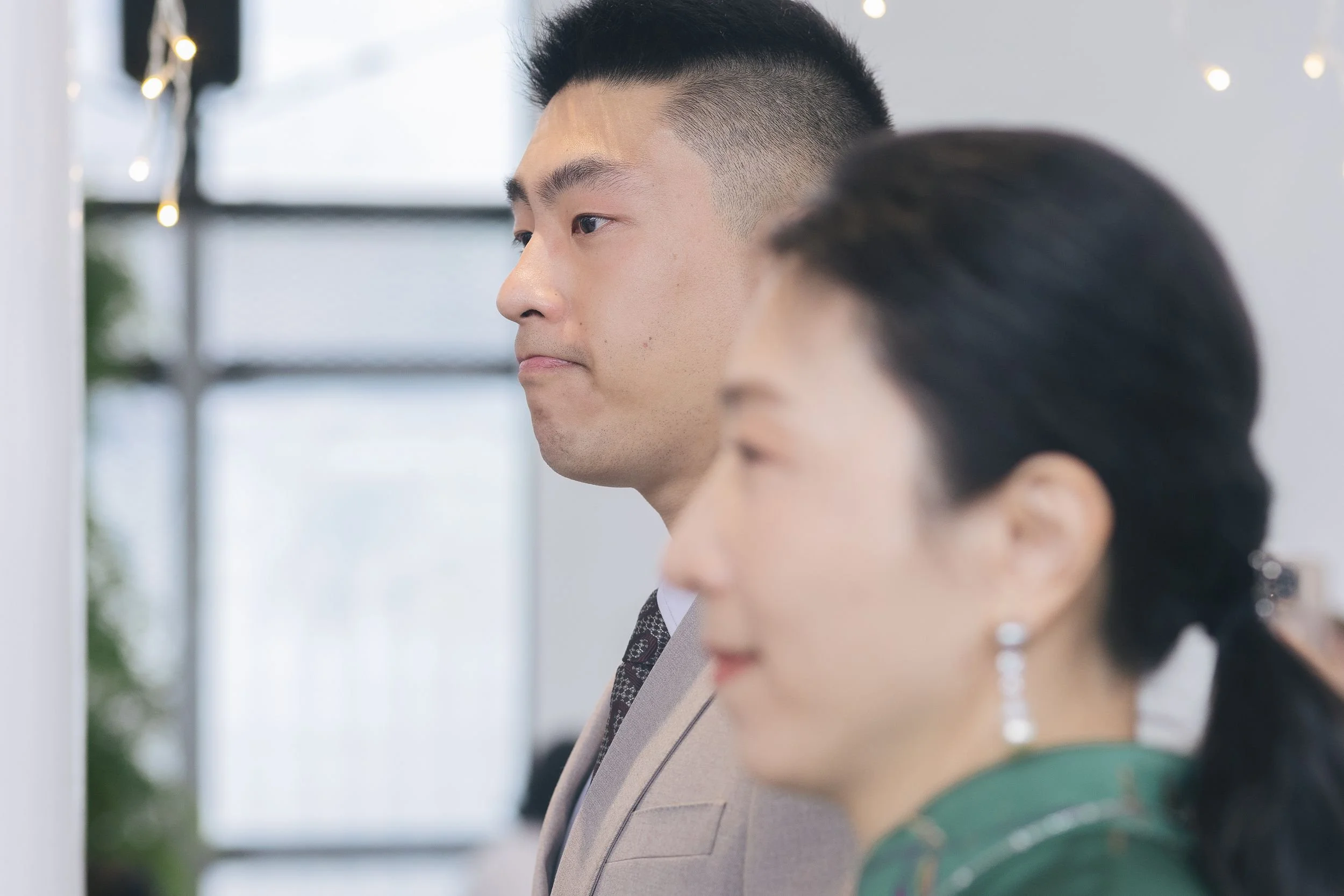 A man with a short, styled haircut and a woman wearing a green top and jewelry are seen in a close-up, side-profile view, standing indoors near a window with blurred background.