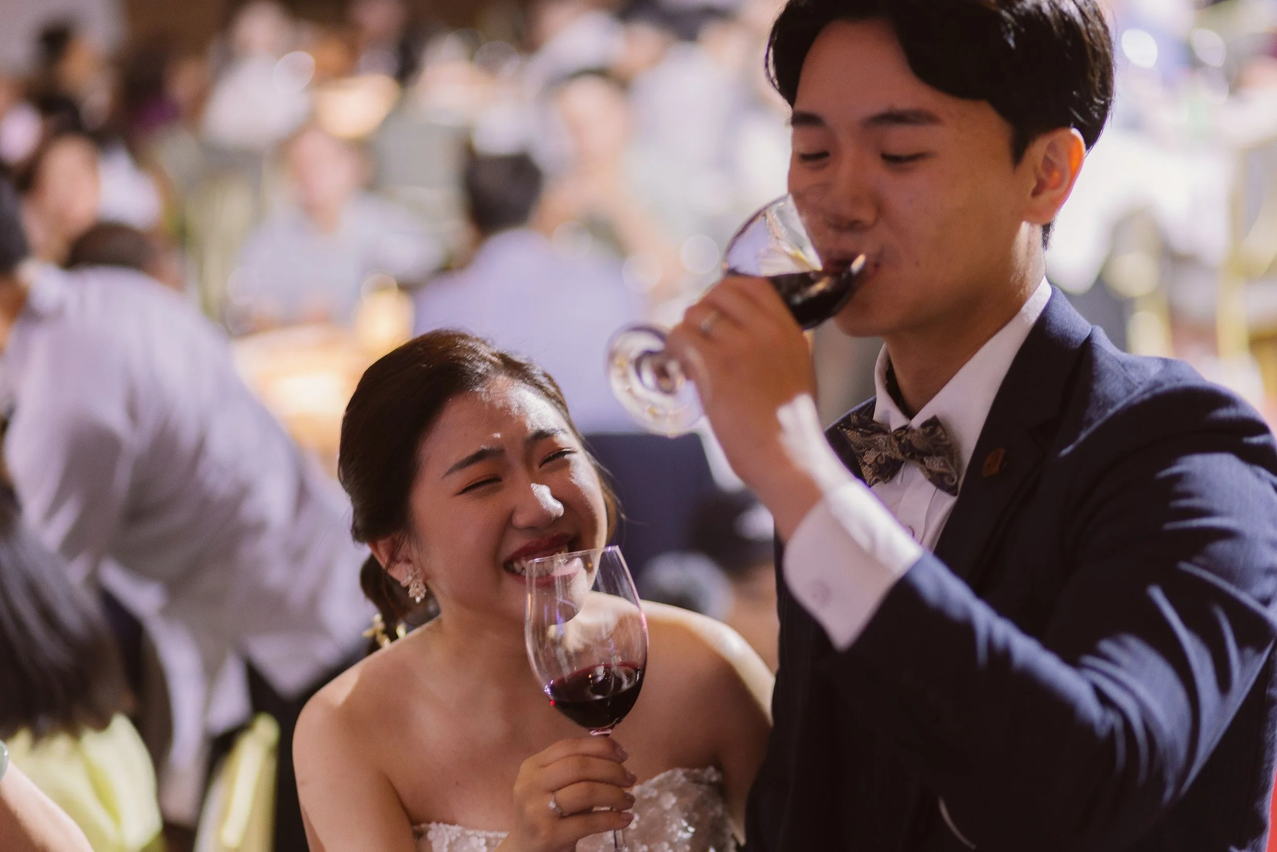 A young woman in a wedding dress smiling and holding a glass of red wine, while a man, dressed in a suit with a bow tie, drinks from his glass. They are at a wedding reception with blurred guests in the background.