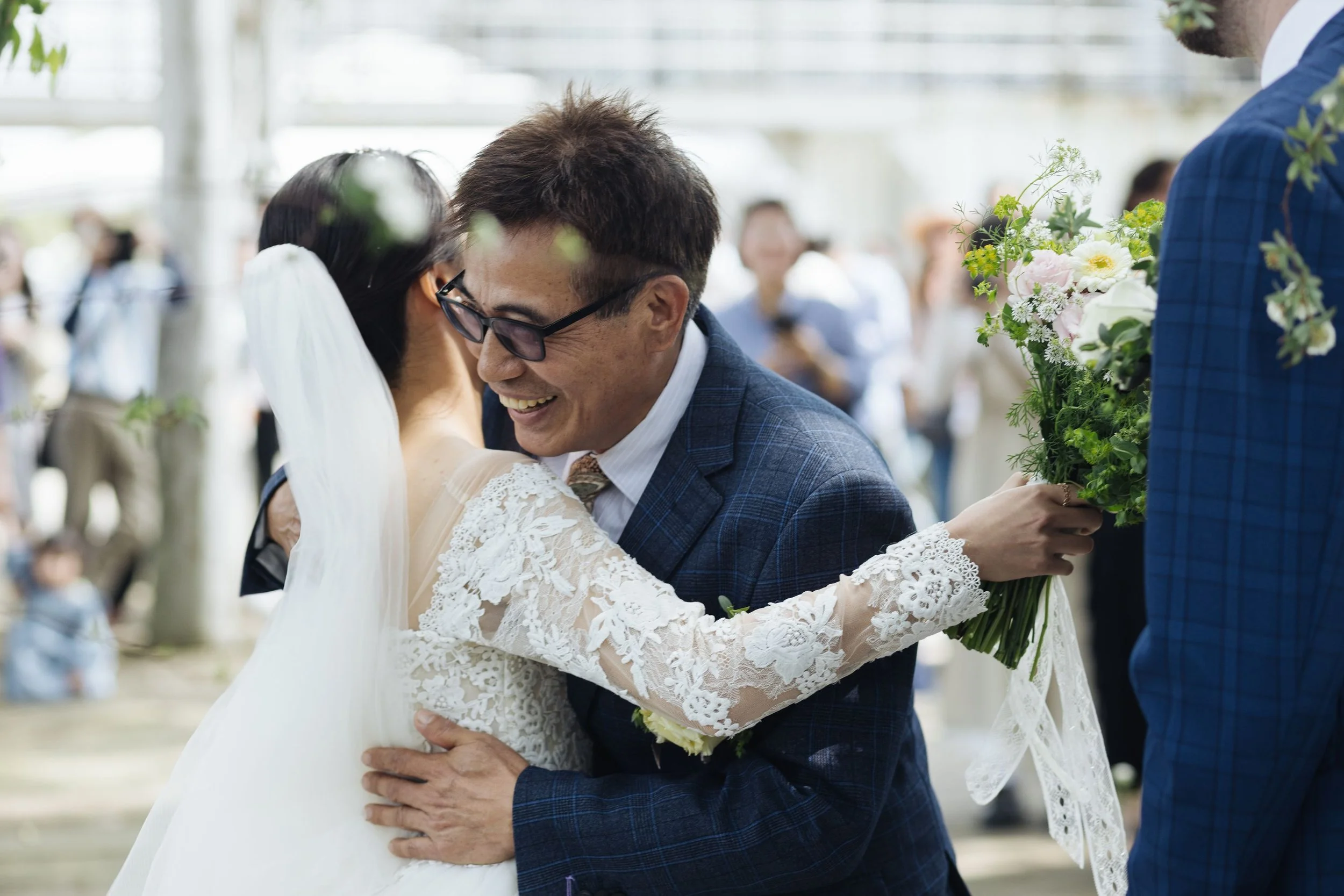 A couple hugging at their wedding with the bride holding a bouquet of flowers, and guests in the background.
