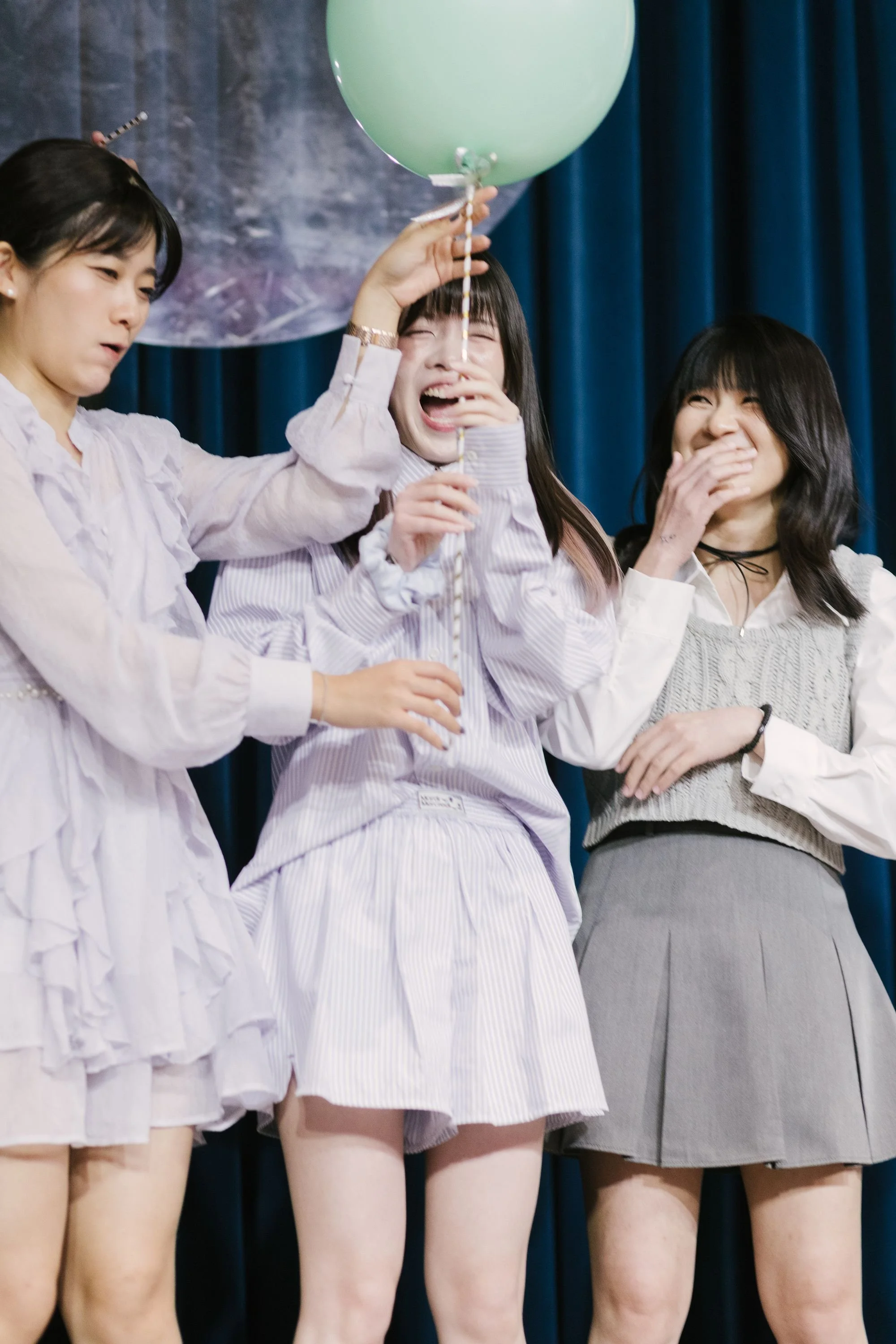 Three young women in skirts and tops standing on stage, one holding a light green balloon, all smiling and laughing.