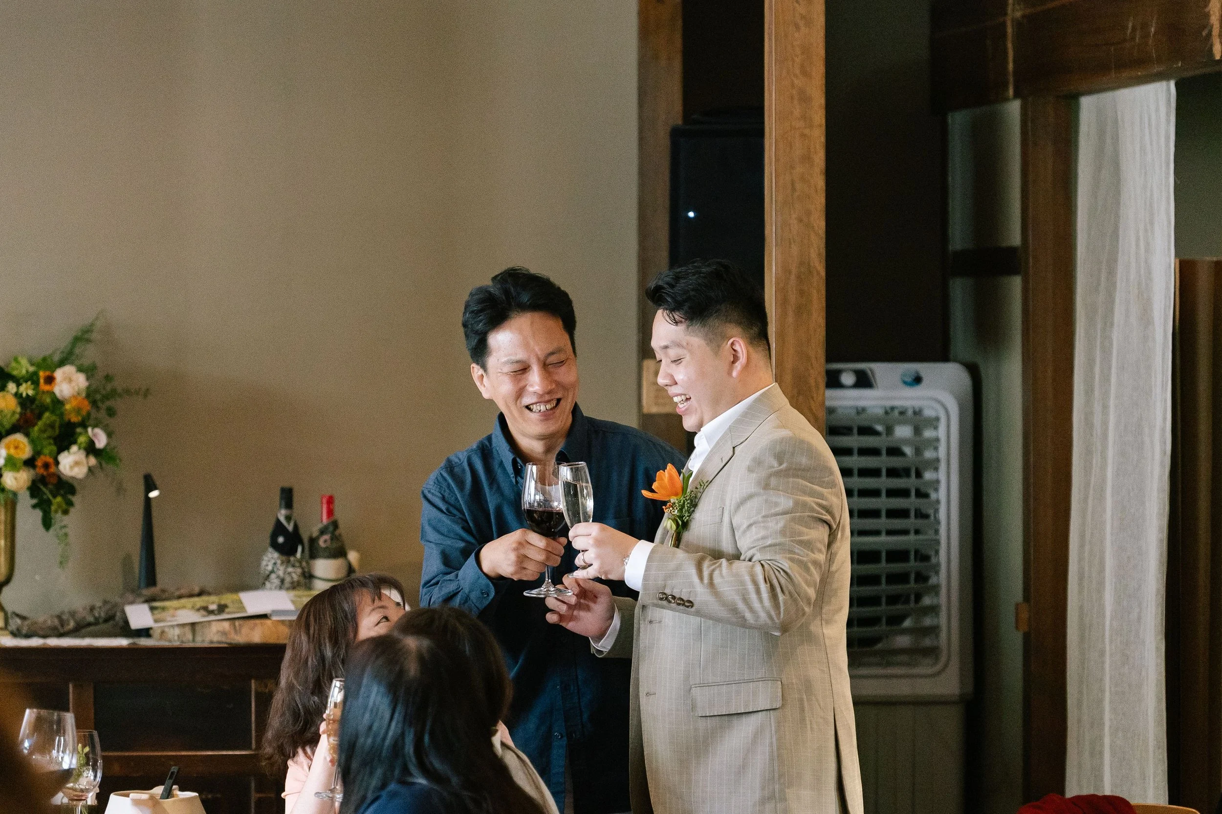 Two men are smiling and clinking glasses of red wine at a celebration, with children seated nearby.