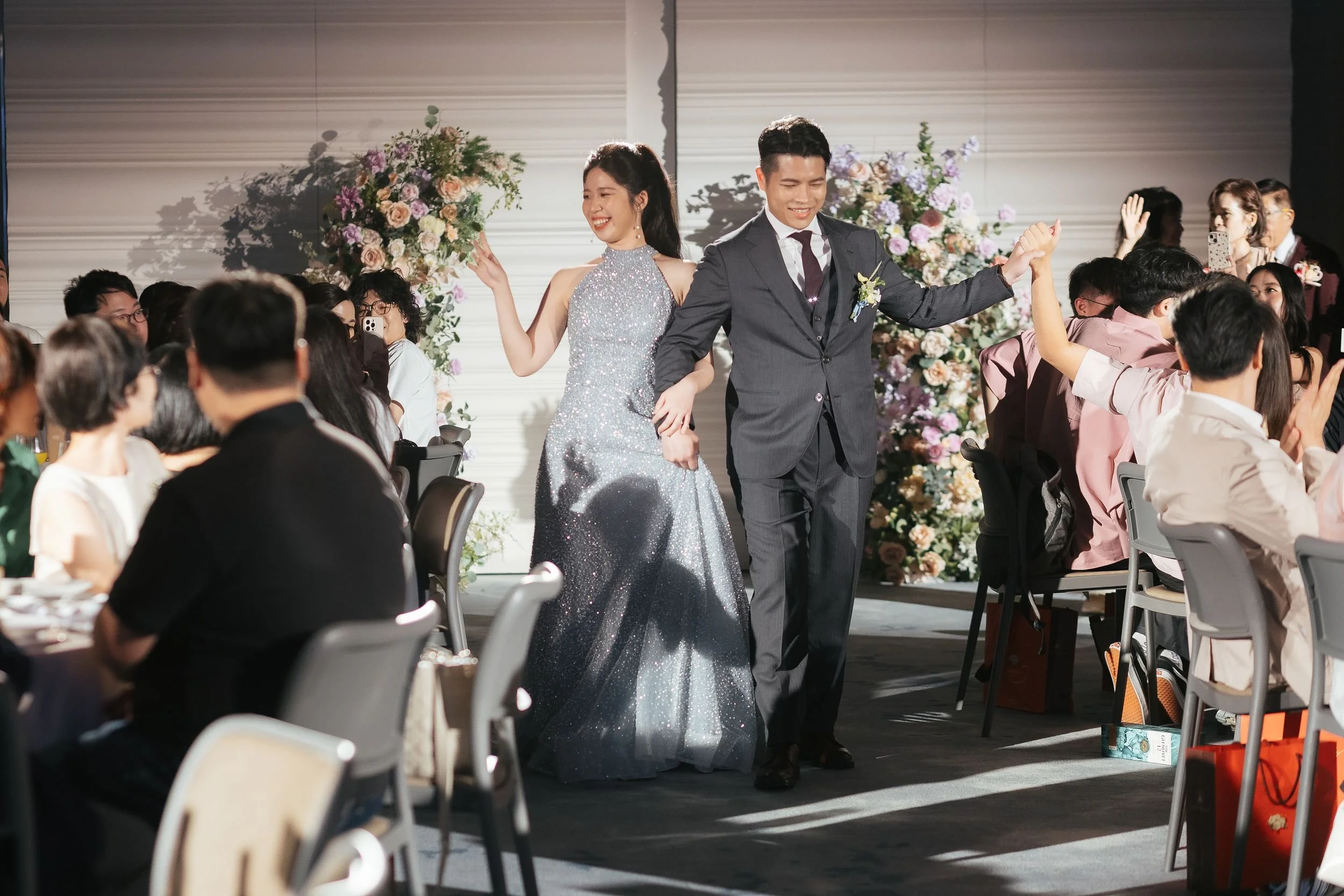 A bride and groom are holding hands and smiling as they walk through a large wedding reception hall filled with seated guests. The bride is wearing a sparkly silver gown and the groom is in a dark suit. The background features floral arrangements and