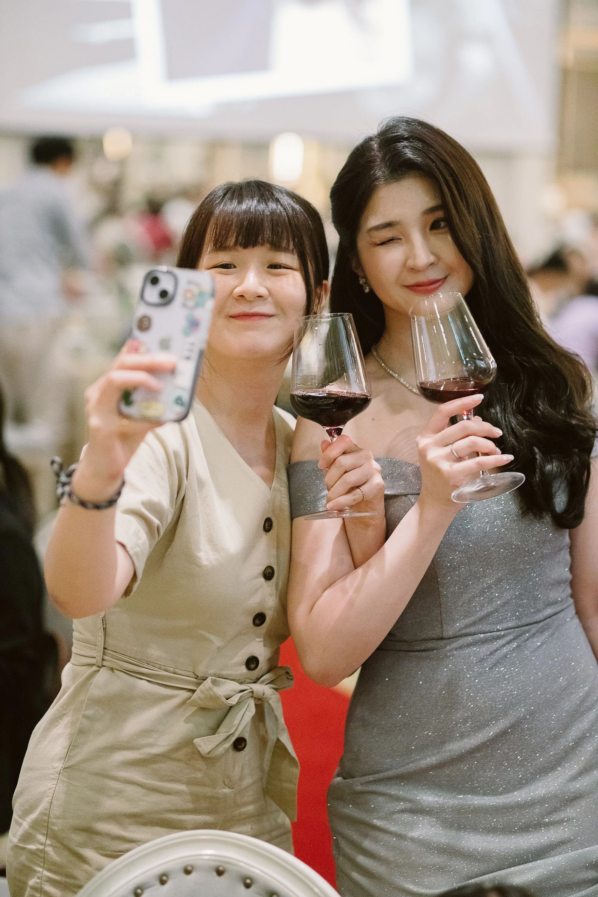 Two women taking a selfie together at a social event, each holding a glass of red wine, smiling and winking at the camera.