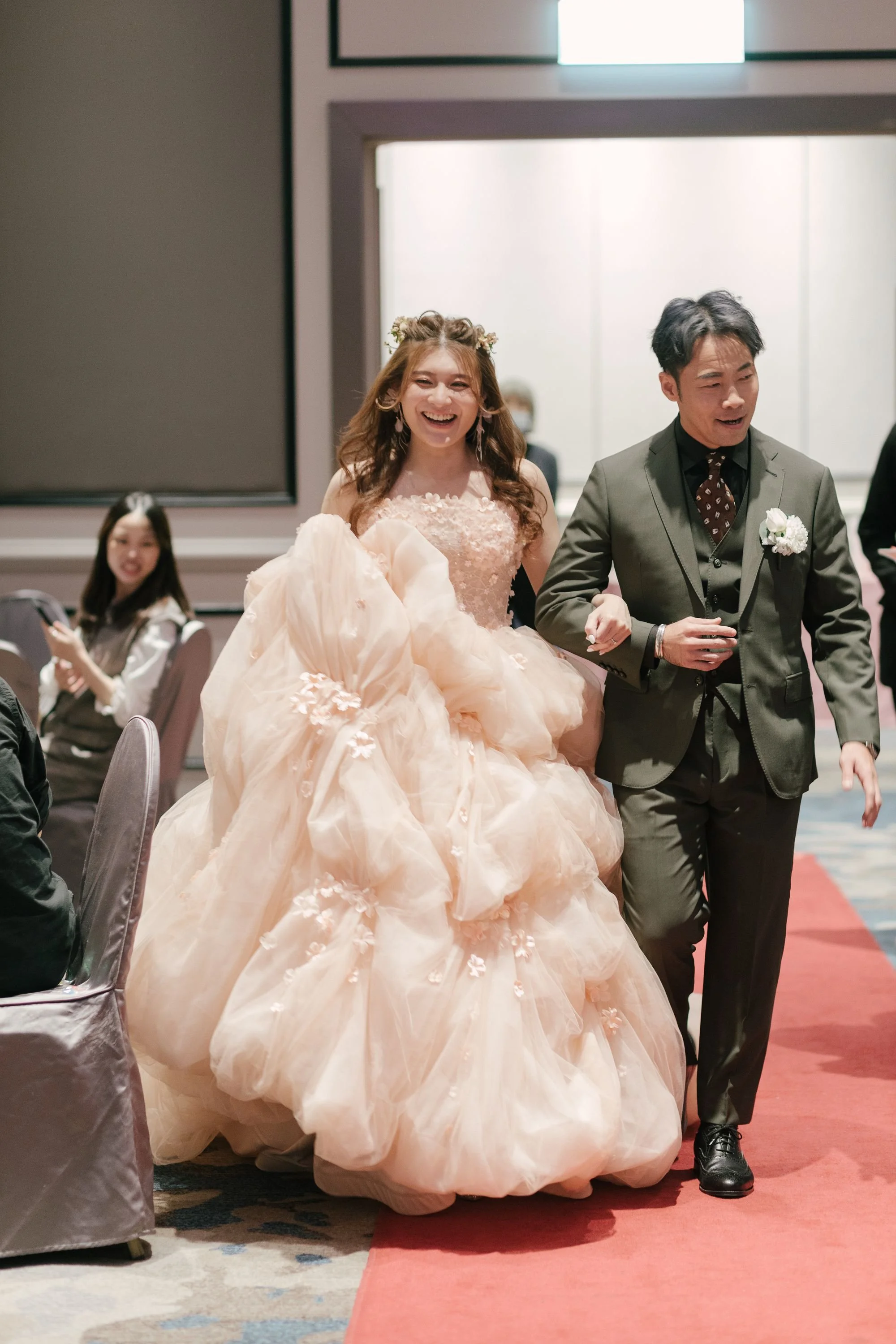A woman in a peach-colored wedding gown with floral details, smiling and holding up her dress, walking hand-in-hand with a man in a dark suit at a wedding reception.