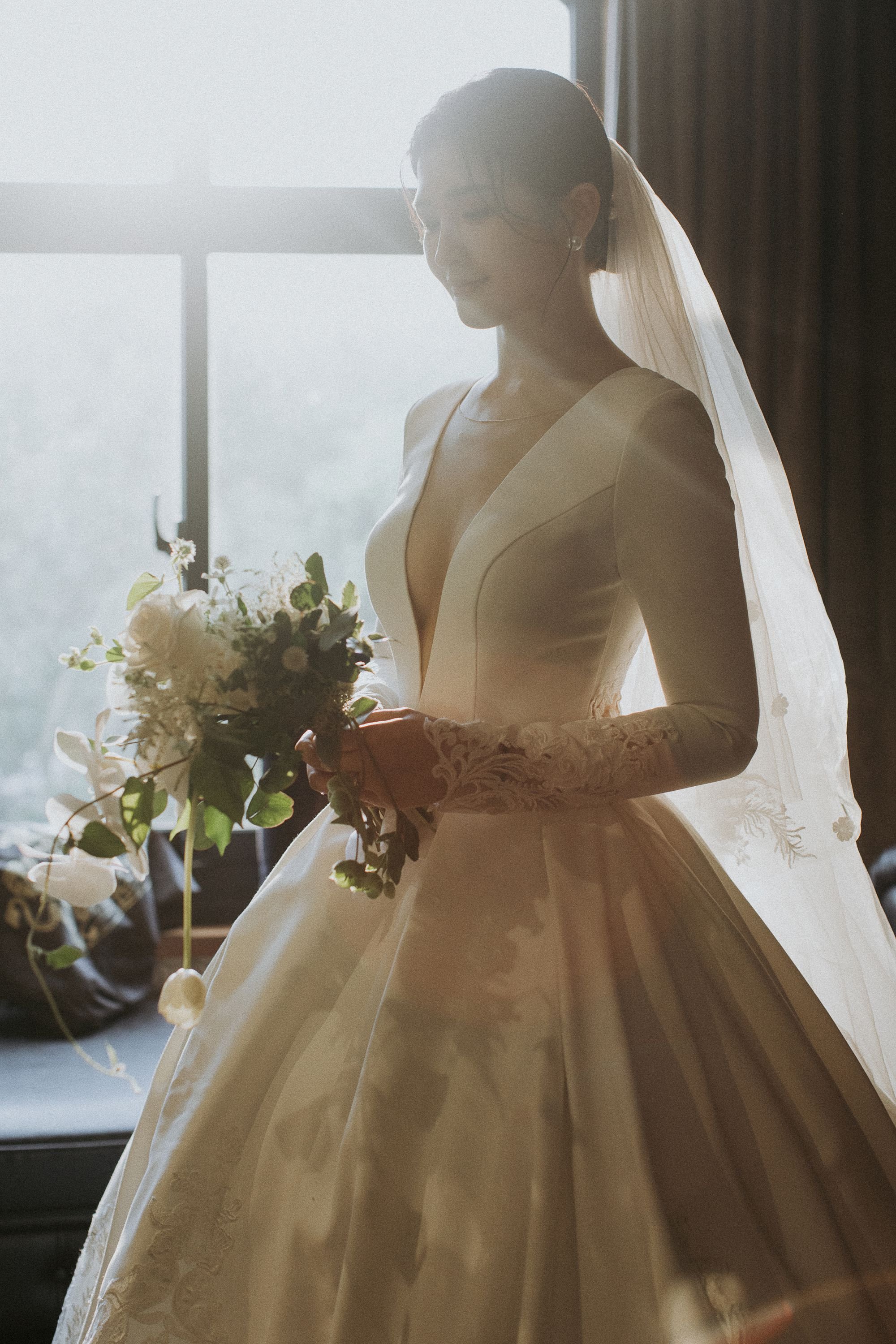 A bride in a white wedding gown and veil, holding a bouquet of flowers, standing by a window with sunlight streaming in.
